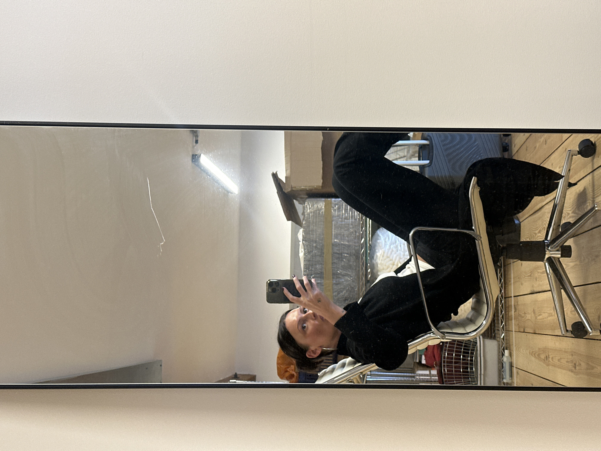 A woman is captured in a mirror selfie, sitting in a light-colored office chair with chrome accents. She holds a dark smartphone, positioning it to photograph her reflection. Her expression is one of mild surprise or playfulness, with wide eyes and a slightly open mouth. She has short, dark hair and is wearing a dark long-sleeved top and dark trousers. Her fingernails are painted with dark polish, and a small tattoo is visible on the back of her left hand.
The mirror takes up the lower portion of the frame, with a plain white wall above it. The reflections in the mirror reveal an indoor setting, possibly an office or studio. To the left, a long fluorescent light fixture is visible on a wall. Behind the woman's reflection, there are metal shelving units holding various items, some appearing to be wrapped in clear plastic or white material. The floor is reflected as light-toned wooden planks. Underneath the chair, a red object, possibly a small fan or bag, and a small bottle are visible. The mirror surface shows some minor smudges and scratches. No discernible text is visible within the image. The general impression is an everyday moment in an indoor, functional space.