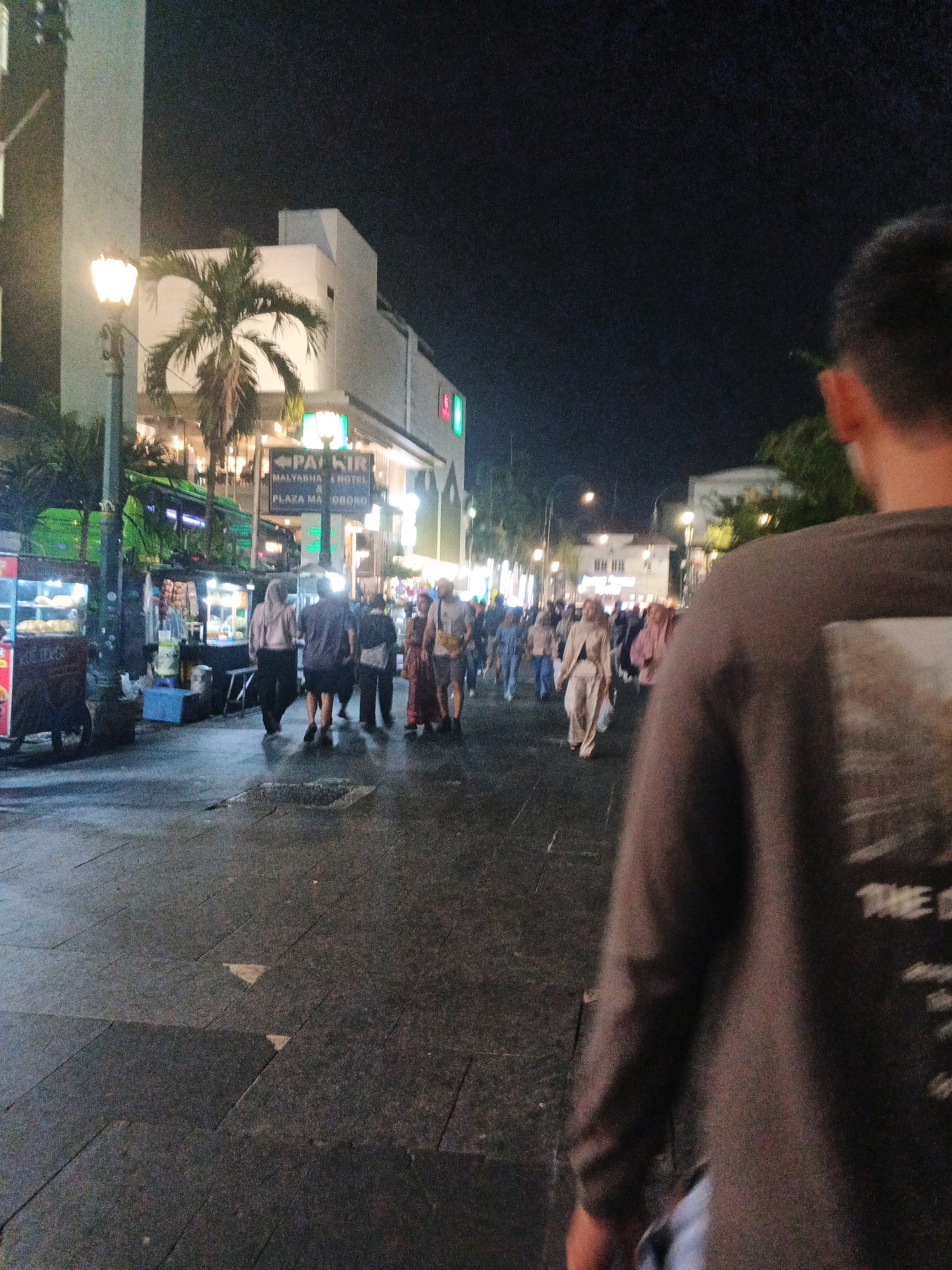 The image depicts a bustling night scene on a city street in Yogyakarta, Indonesia. The perspective is from behind a person, with their back to the camera, wearing a dark long-sleeved shirt with a graphic on the back that reads "THE D". The street is paved with dark rectangular tiles, and there are puddles of water reflecting the surrounding lights.
To the left of the frame, there are several food stalls with illuminated displays showcasing food items, likely street food vendors. One stall has a glass-fronted display case filled with what appear to be bread rolls. Streetlights cast a warm glow on the scene.
Further down the street, a group of people are walking. Many are dressed in casual clothing, and some women are wearing headscarves, indicating a likely Indonesian setting. In the background, buildings are visible, including what appears to be a hotel with a prominent "PARKIR" (parking) sign pointing towards "MALYARUHA HOTEL" and "PLAZA MALIOBORO." A palm tree is situated in front of one of the buildings. The overall atmosphere is lively and suggests a popular area for pedestrians and dining. The image is characterized by its nighttime lighting and the presence of a crowd.
