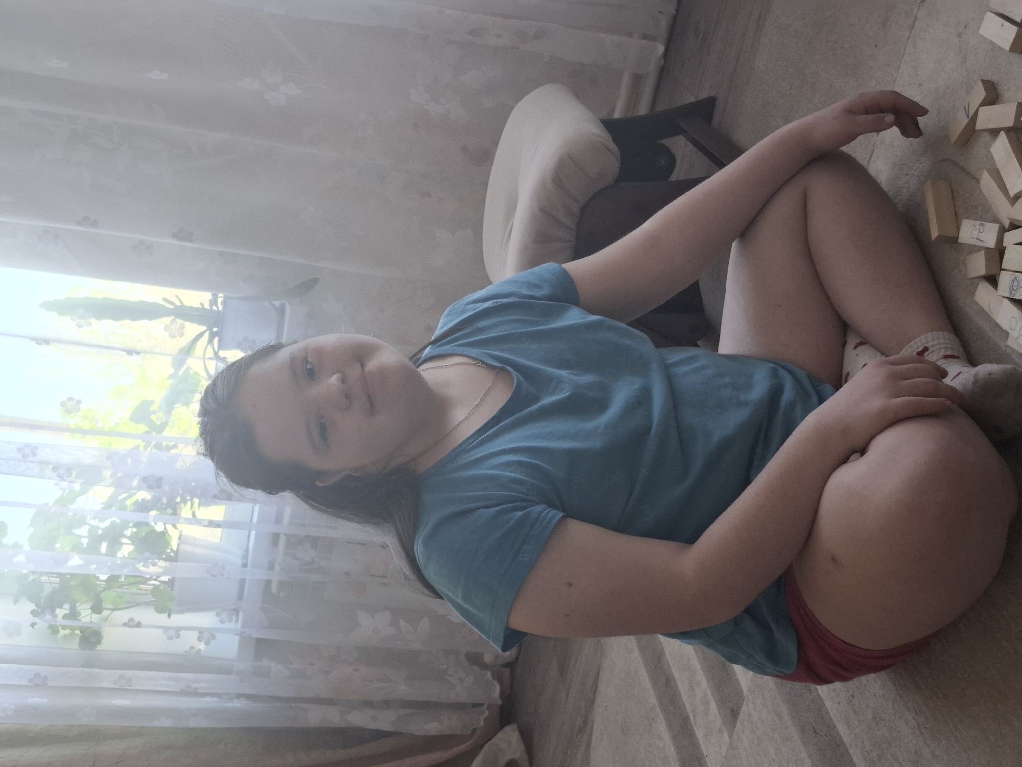 A young woman is sitting cross-legged on the floor of a room. She wears a blue t-shirt and red shorts and patterned socks. A small wooden chair is to her left. A pile of wooden blocks with writing on them is on the floor near her. A window with sheer curtains is behind her, with greenery visible outside. The flooring appears to be beige carpet. Without additional location specific details, it is not possible to confirm it is Chaikovsky, Russia.