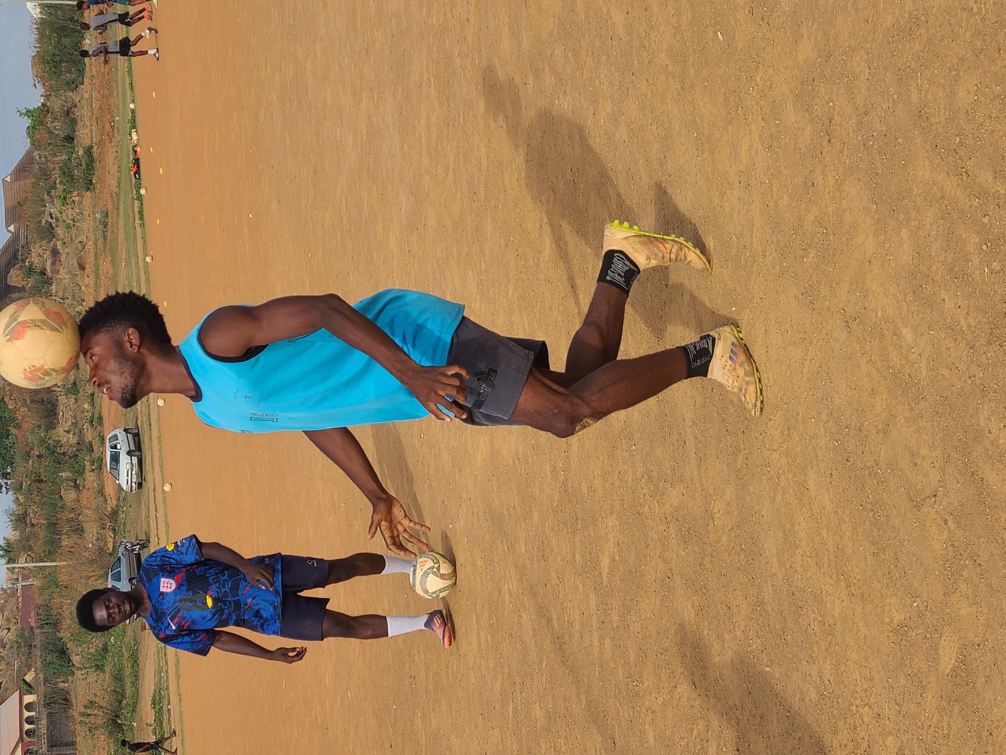 The image depicts two young men engaged in a soccer activity on a dirt field in Barikin Jirga Summa, Nigeria. One man, wearing a bright blue sleeveless shirt and dark shorts, balances a soccer ball on his forehead. The other man, wearing a blue soccer jersey with an England flag and dark shorts, is juggling a soccer ball with his hand. The setting includes dry, brownish ground, some distant buildings, vegetation, and vehicles in the background. Several other soccer balls are also visible on the field.