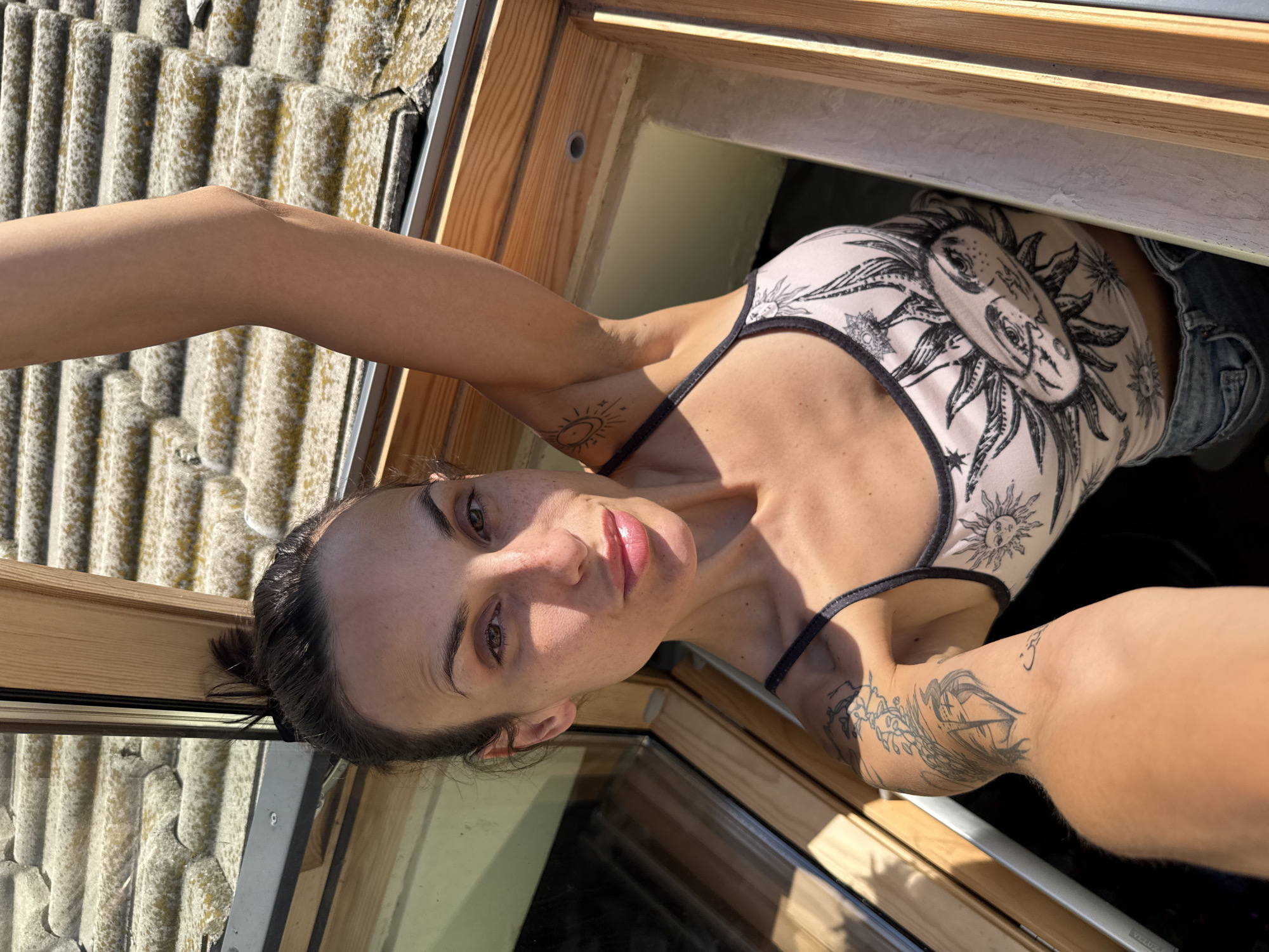 A woman with tattoos and dark hair pulled back is taking a selfie while leaning out of a skylight. She is wearing a light-colored tank top with a sun and moon design and appears to be wearing denim shorts. Her left arm is extended above her head, and her right arm is holding the camera. The roof outside the skylight is visible and covered in gray shingles. The interior of the skylight frame appears to be natural wood. Based on the direction of the light, it is likely daytime. The location is Pleven, Bulgaria is not identifiable from this image.