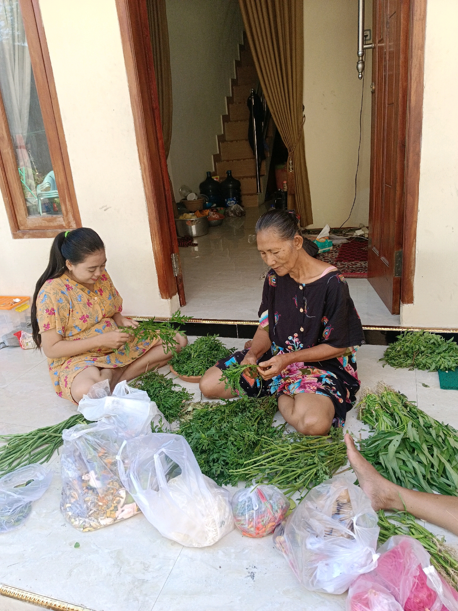 Two Indonesian women, an older woman and a younger woman, are sitting on a tiled floor in what appears to be a covered outdoor area or a porch of a house. They are both engaged in processing leafy green vegetables. The younger woman, wearing a yellow floral print dress, has her hair tied back in a ponytail and is looking down at the greens she is holding. The older woman, with graying hair and wearing a black top with colorful patterns, is also focused on the vegetables. Both women are surrounded by large piles of these greens, some of which are bundled and tied. Several plastic bags filled with various items, including what appear to be other vegetables and possibly food ingredients, are scattered on the floor around them. In the background, a doorway leads to an interior space with stairs and some large water bottles. The overall scene suggests a domestic activity related to food preparation or market sales. The location is identified as Krembangan, Indonesia.