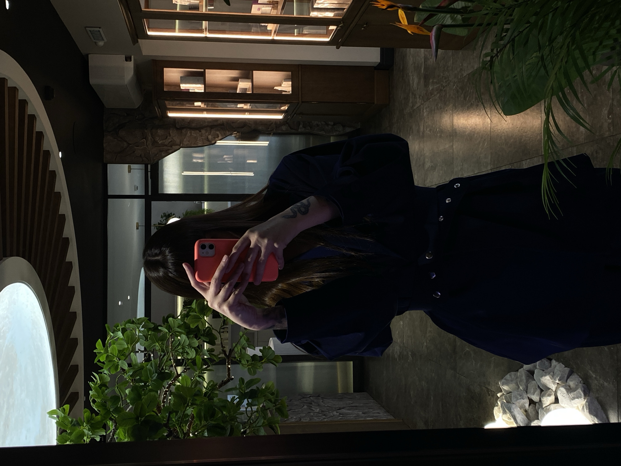The image depicts a person taking a mirror selfie in what appears to be an upscale indoor commercial space, possibly a cafe, restaurant, or retail store, located in Votkinsk, Russia. The image is oriented sideways, but the description assumes an upright perspective.
The central subject is a person, likely female, with long, dark hair, holding a coral-colored smartphone with both hands, obscuring her face as she captures her reflection. A tattoo, appearing to be a snake or dragon, is visible on her right forearm. She is wearing a dark, possibly navy blue or black, long-sleeved dress or coat with silver buttons down the front.
The setting is characterized by a blend of modern and natural design elements. To the left, a large, glowing white circular or semicircular light fixture provides a striking visual. In the foreground, a vibrant green potted plant adds a touch of nature. The background features a distinctive wall section crafted from stacked, light-colored artificial rocks, illuminated from below to create a dramatic effect. Further behind, display shelves with warm strip lighting showcase various items, possibly books or merchandise. Another large, leafy green plant with bright yellow-orange flowers (resembling a Bird of Paradise) is visible on the right. The floor is dark and reflective, mirroring some of the ambient light. Architectural details include dark walls, glass partitions, a ceiling-mounted air conditioning unit, and a sensor.
The activity is a self-portrait taken via a mirror. The time of day appears to be evening or night, indicated by the reliance on artificial lighting and the dark ambiance visible through what might be windows in the background. No discernible text is visible within the image.