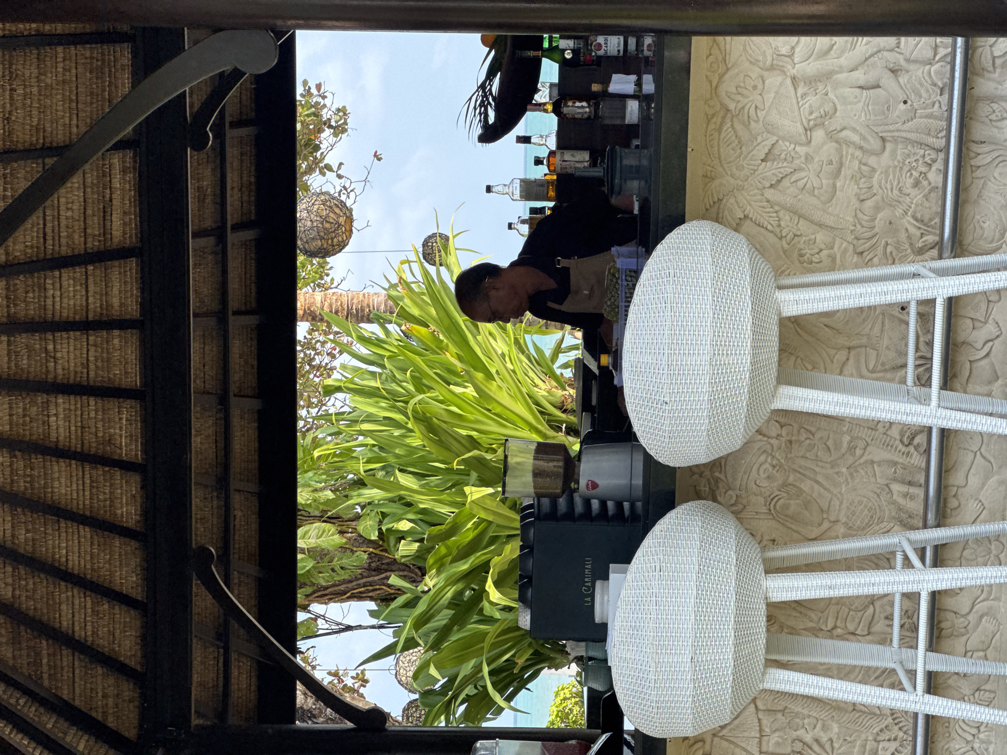 The image depicts an open-air bar at what appears to be a resort or hotel in Kuta Selatan, Indonesia. Two white woven bar stools are positioned in the foreground, facing a bar counter. Behind the counter, a man wearing an apron is preparing drinks, with various bottles of alcohol and a coffee grinder visible on the shelf. Lush green foliage is abundant behind the bar, suggesting a tropical setting. In the background, the turquoise ocean and a cloudy sky are visible. The bar structure itself is thatched, and decorative woven lanterns hang from the ceiling. The base of the bar features intricate carved relief panels, possibly depicting local motifs.