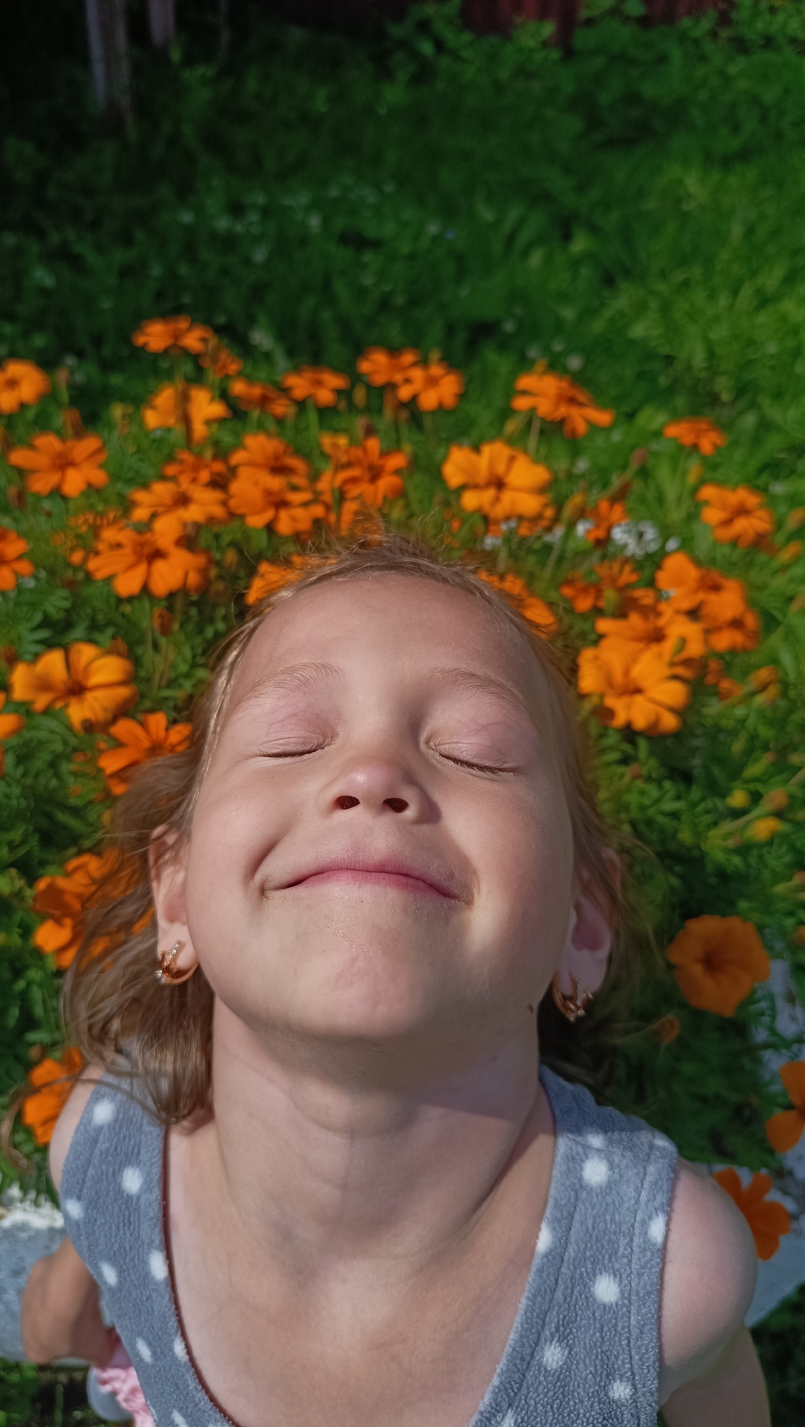 A close-up, high-angle shot shows a young Caucasian girl with blonde hair lying down outdoors. Her eyes are closed, and she has a contented smile on her face. She is wearing a gray, polka-dotted sleeveless top. Small hoop earrings are visible in her ears. The background is filled with green foliage and numerous bright orange marigold flowers. The scene appears to be a garden or park.