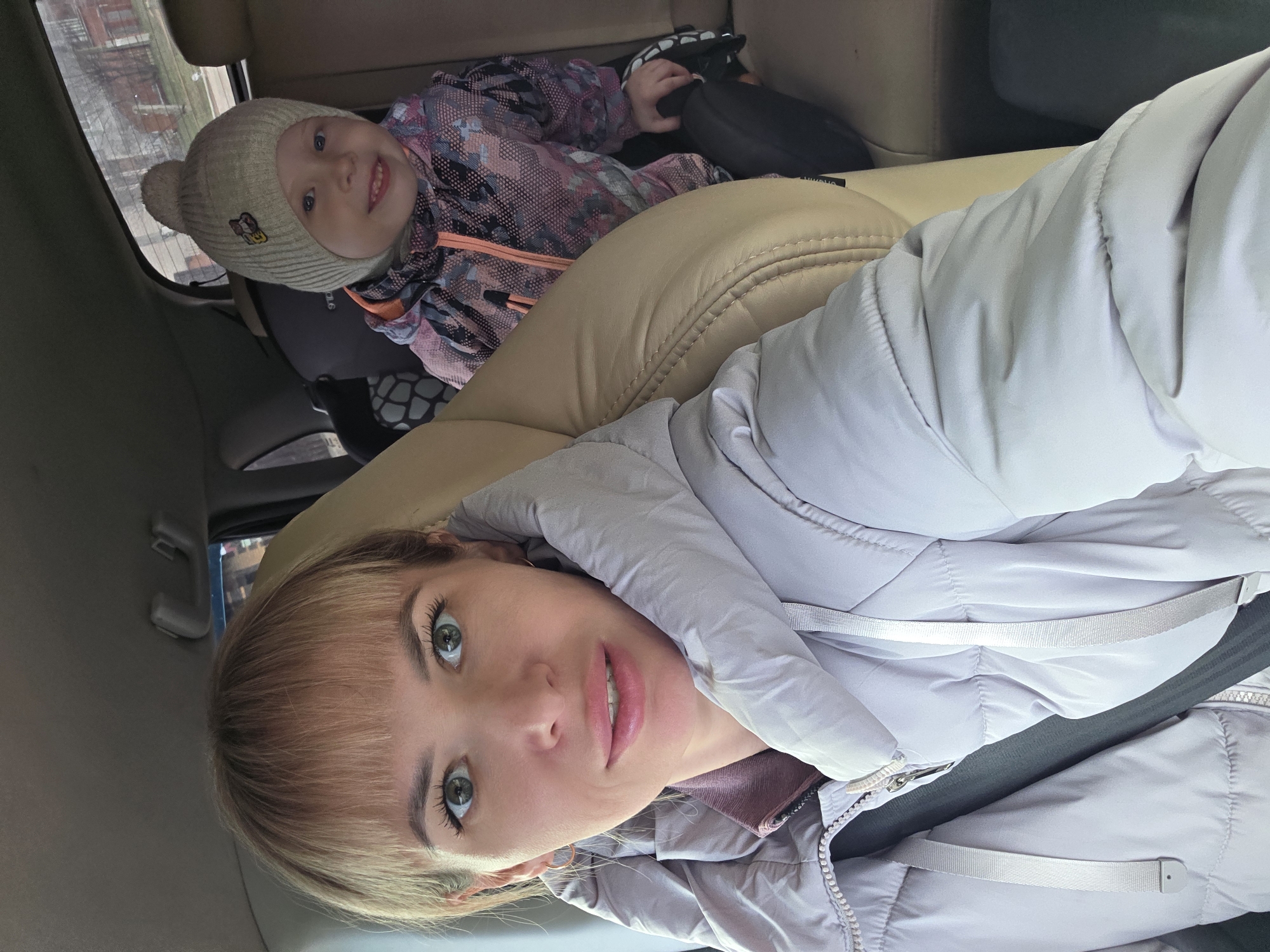 A woman and a young child are captured in a selfie taken inside a car. The image is oriented vertically, suggesting it was taken from a smartphone.
The woman, in the foreground, has blonde hair with bangs and striking light blue/green eyes. She wears a light lavender or grey puffer jacket over a darker top and has a neutral, direct gaze towards the camera. Her facial expression is calm. A car seatbelt is visible across her chest.
In the background, on the right side of the frame, a young child with fair hair and blue eyes smiles brightly at the camera. The child wears a light beige ribbed beanie with a small, colorful cartoon emblem (possibly Minnie Mouse) and a patterned winter jacket in shades of pink, orange, black, and white. The child is seated in a car seat, partially visible.
The interior of the car features light tan leather seats. Through the car window in the upper left background, indistinct residential buildings and what appears to be an overcast sky can be seen, suggesting daytime and cool weather, consistent with the subjects' winter attire. No specific text is clearly legible in the image, apart from the small emblem on the child's hat. The overall mood is one of contentment, possibly during a car journey or outing in Chaikovsky, Russia.