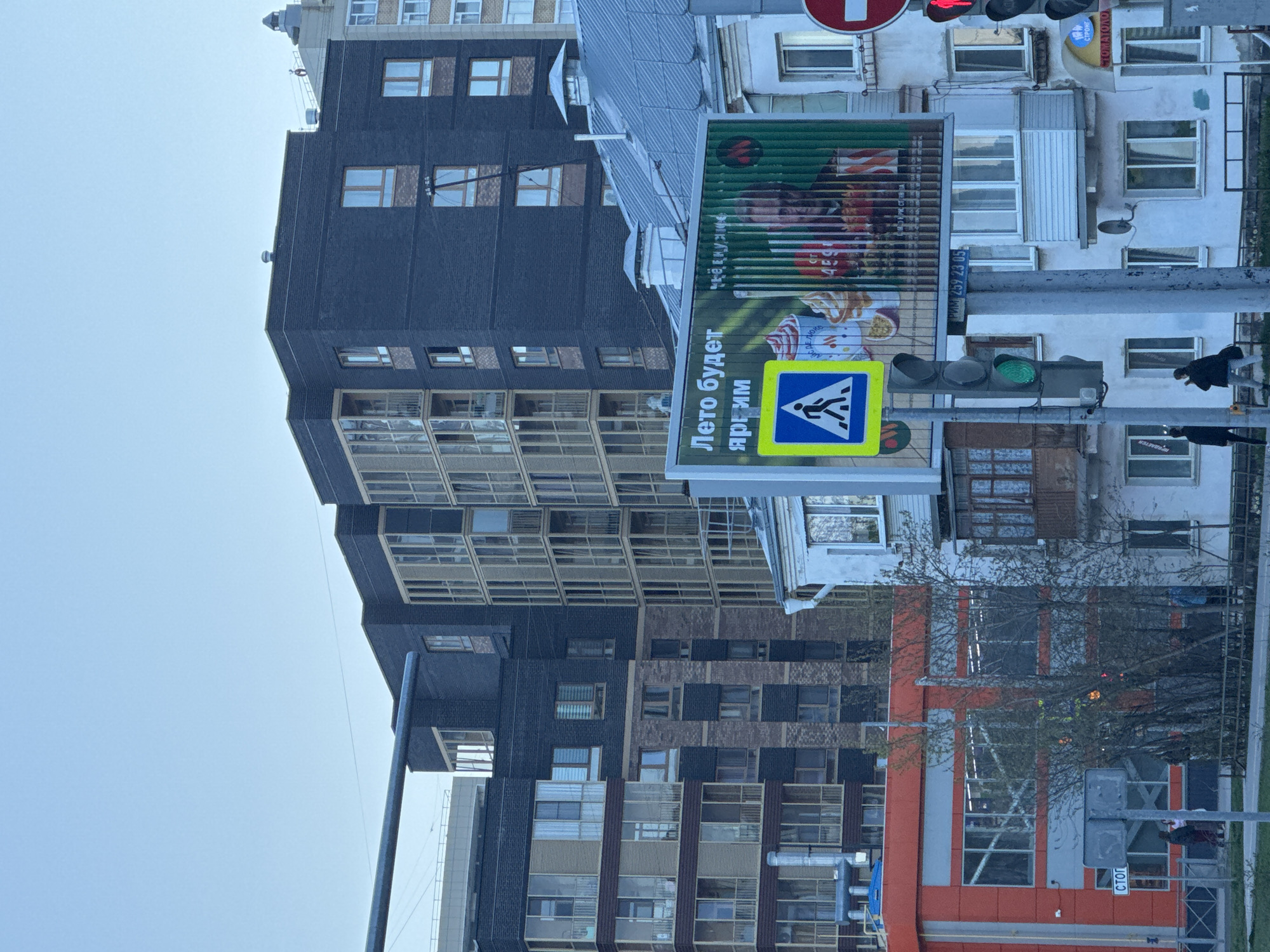 The image shows an urban scene in Perm, Russia. Buildings of varying heights and architectural styles line the street. A pedestrian crossing sign with the text "Лето будет ярким" (Summer will be bright) in Russian is visible. Traffic lights are also present, with the green light illuminated. Pedestrians are standing by the traffic light, waiting to cross the street. A billboard advertising products is affixed near the pedestrian crossing sign. The sky is overcast. The image is taken in daylight.