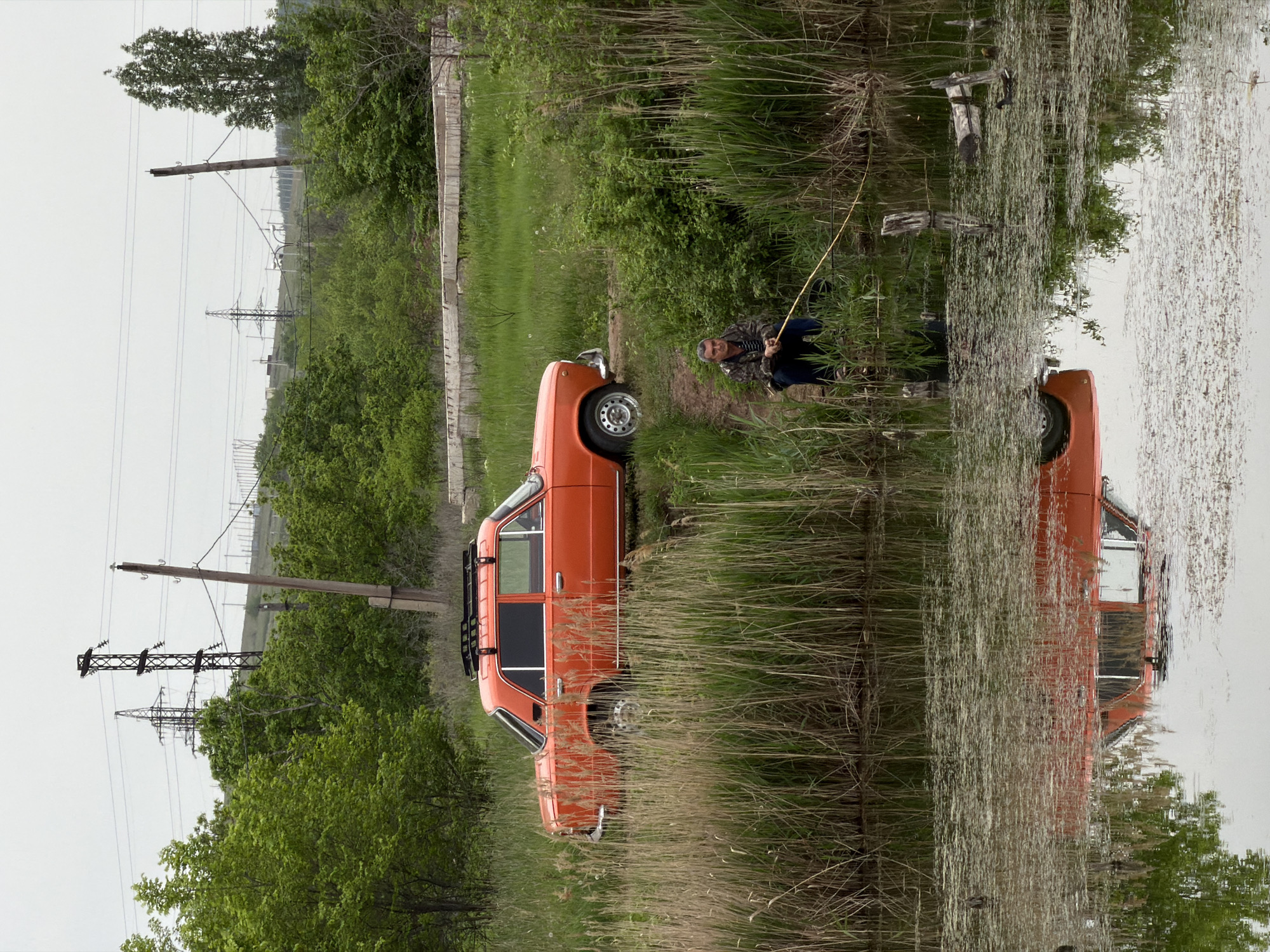 The image depicts a man fishing in a body of water next to an orange vehicle parked on the bank. Reeds and vegetation grow along the water's edge, with the vehicle's reflection visible in the water. In the background, power lines and green trees are visible. The location is Kamyshin, Russia.