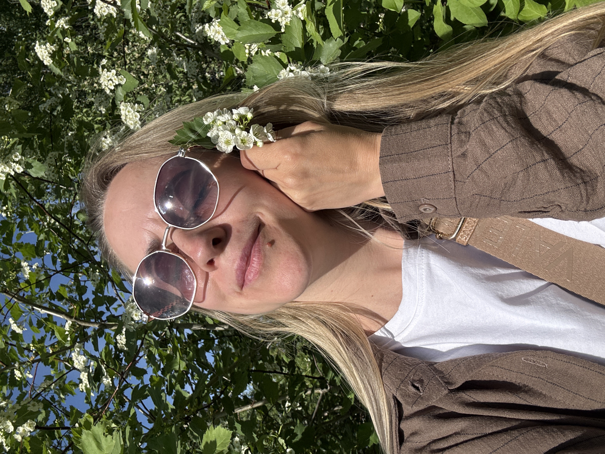 A woman is posing outdoors in Chaikovsky, Russia. She has long blonde hair and is wearing sunglasses, a white t-shirt, and a brown shirt. She has flowers tucked behind her ear. The background includes foliage and trees with white blossoms, some of which are in focus and some are blurred. A shoulder bag strap is visible across her chest. The scene is brightly lit, suggesting it is daytime.