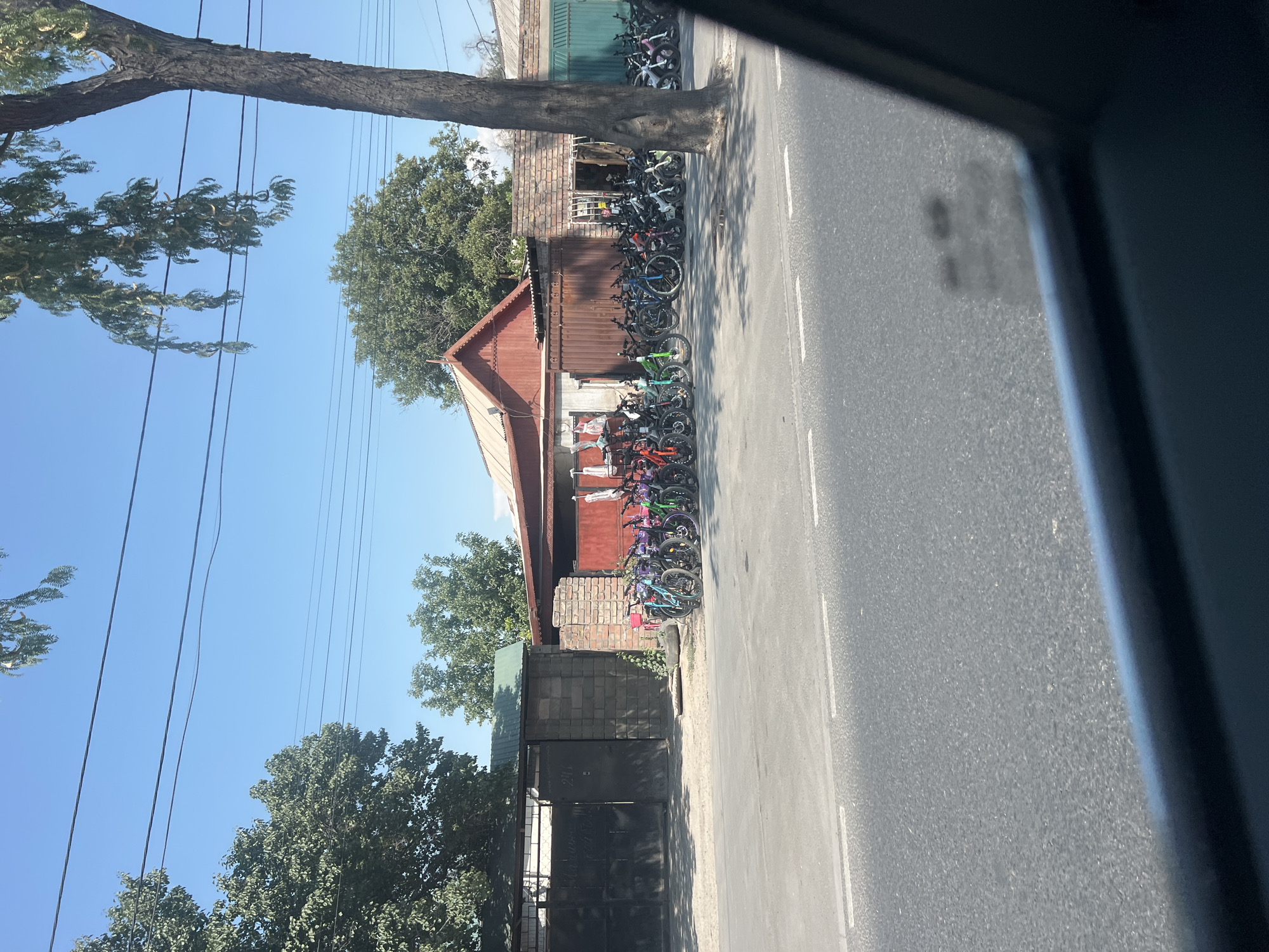 In the city of Bishkek, Kyrgyzstan, on a sunny day, a row of bicycles is parked along a street in front of a building. The bicycles are of various colors and sizes, and some appear to be electric bikes. The building behind the bicycles is a single-story structure with a red roof and brick walls. A large metal gate is visible to the left of the building, and trees are scattered throughout the scene. The street in front of the building is paved and has white lane markings. The overall impression is one of a commercial area, possibly a bicycle shop or rental service.