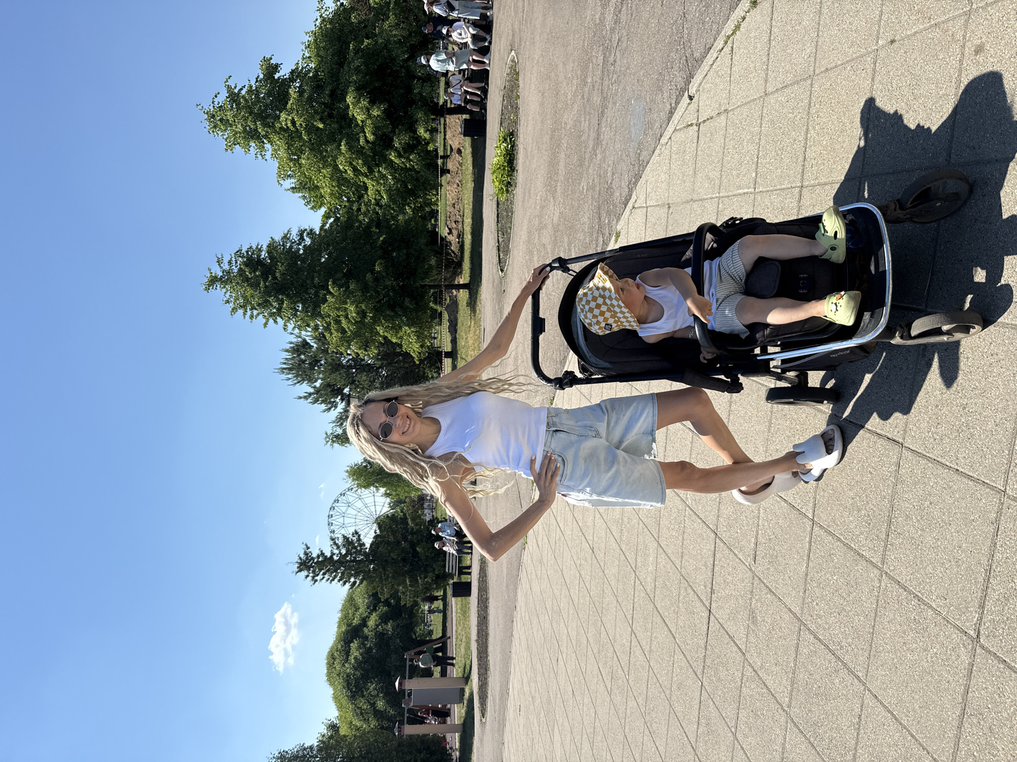 The image depicts a sunny outdoor scene in the city of Zavolzhye, Russia. In the foreground, a woman with long blonde dreadlocks and sunglasses stands next to a stroller. She is wearing a white tank top and light blue denim shorts, with her left hand on her hip and her right hand resting on the stroller. Inside the stroller, a young child wearing a checkered hat and striped shorts is seated.
The background reveals a paved walkway with a tiled pattern, bordered by green grass and trees. Further in the distance, several people are visible sitting on benches, and a playground area with swings can be seen. A Ferris wheel is partially visible behind the trees, suggesting the presence of an amusement park or recreational area. The sky is clear and blue, indicating fair weather.