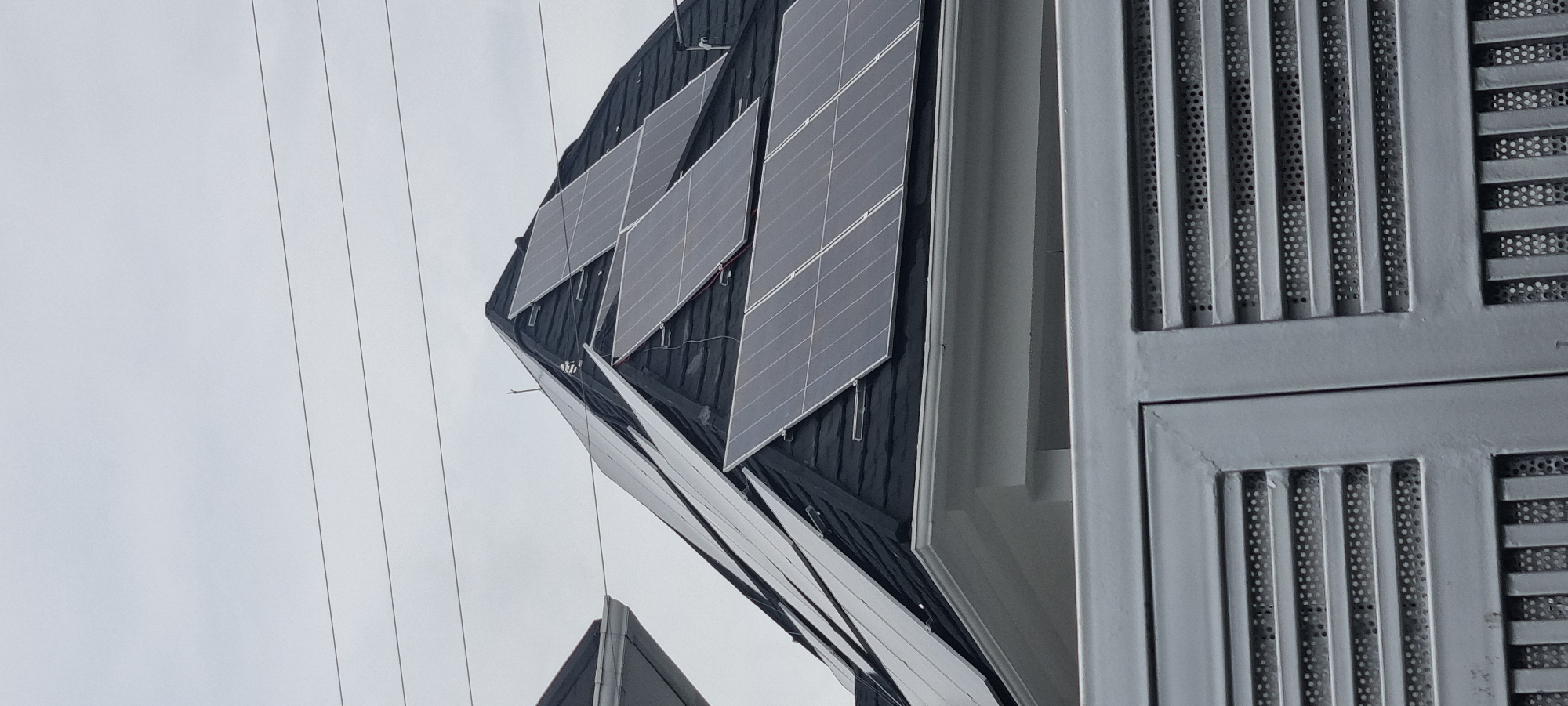 The image shows a close-up of solar panels installed on a dark-tiled roof of a building in Moba, Nigeria. The sky is overcast and appears grayish. Several solar panels are visible, arranged in rows and angled towards the sky. Some panels are a darker shade of gray, while others appear lighter, possibly due to reflections or different types of panels. There are visible wires and mounting hardware attached to the panels and the roof. In the foreground, a portion of a light gray metal fence with horizontal slats and a perforated pattern is visible, partially obscuring the lower part of the building. The overall scene suggests a residential or commercial building equipped with renewable energy technology.