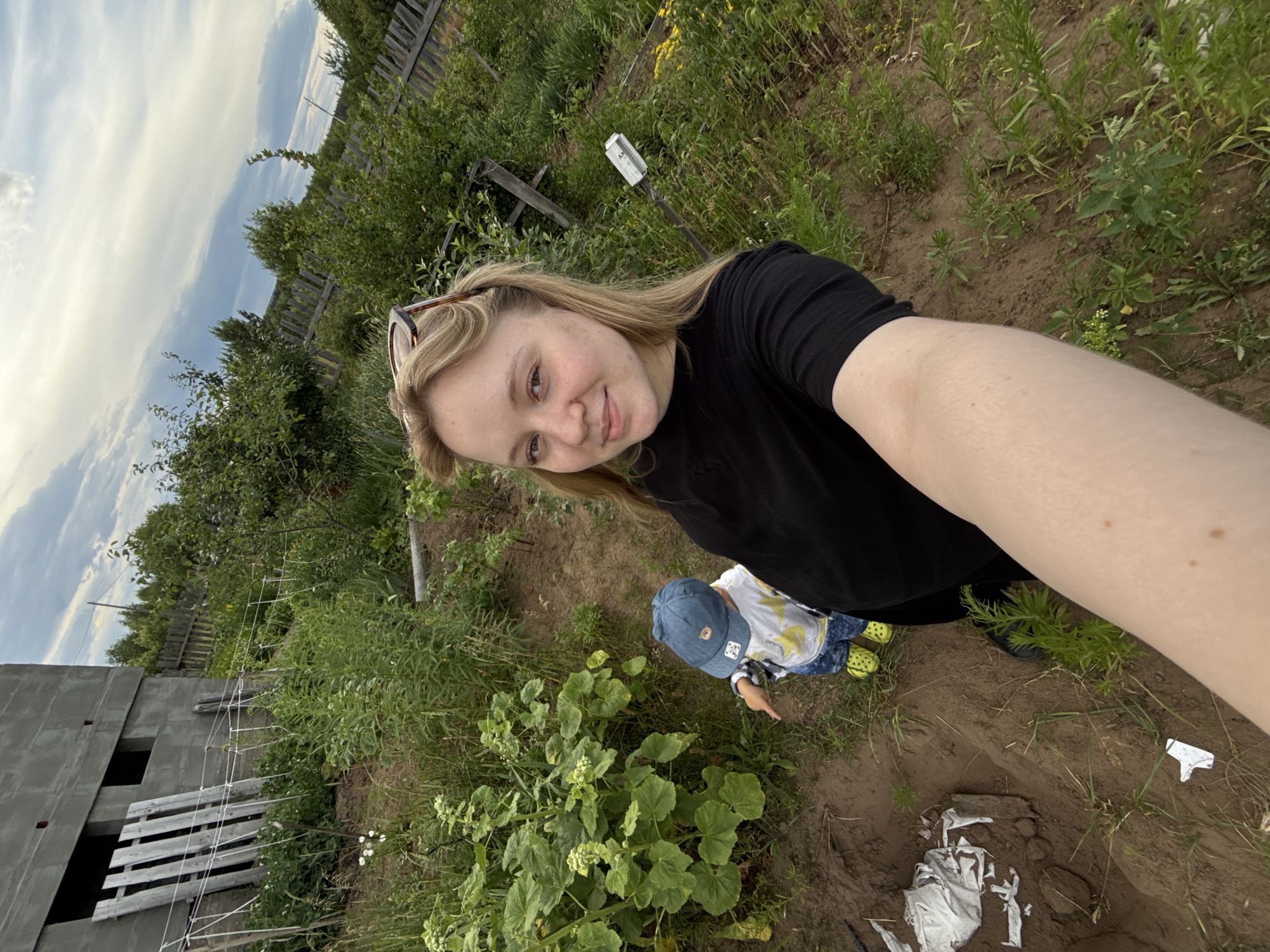 A woman and a young child are in an outdoor scene in Гаревая, Russia. The woman is taking a selfie, holding the camera with her right arm. She has blonde hair and is smiling at the camera. She is wearing a black t-shirt and has sunglasses perched on her head. The child, facing away from the camera, is wearing a blue cap, a white and yellow patterned shirt, and bright yellow shoes. The child is pointing at a large leafy plant.
The setting appears to be a backyard or garden area. In the background, there is a partially constructed building made of concrete blocks. Further in the distance, there is a wooden fence and lush green vegetation, including trees and tall grass. The ground is a mix of dirt and some sparse vegetation. Some white cloth is scattered on the ground near the woman's feet.
The sky is visible with some clouds, suggesting it could be daytime, possibly late afternoon or early evening given the soft light. The overall mood seems relaxed and casual, with the woman appearing happy. There is no visible text in the image.
