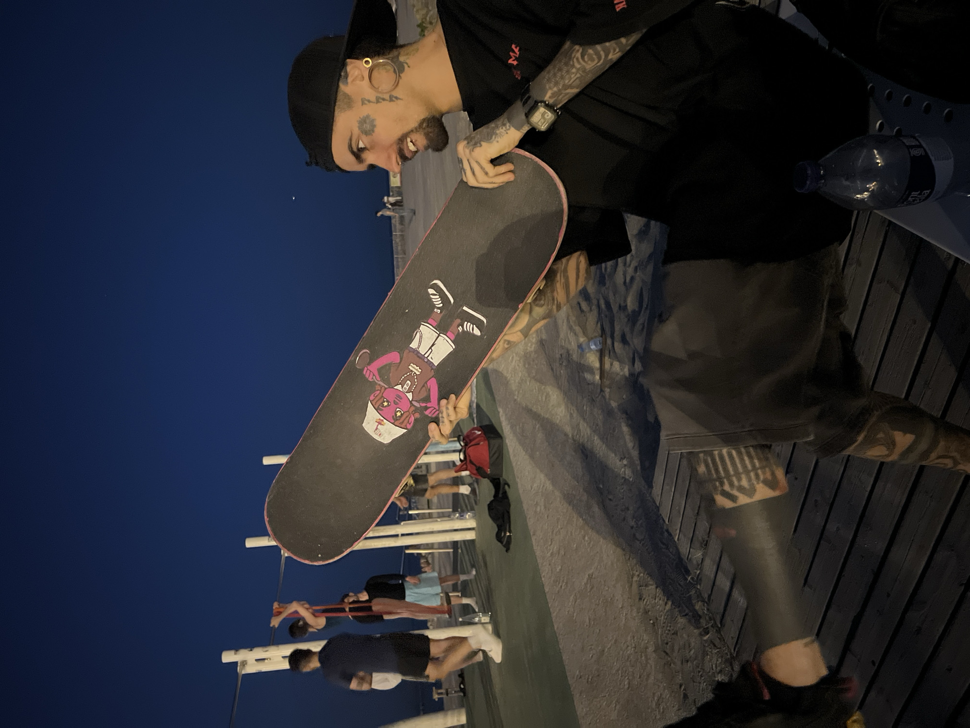 A person stands outdoors at night, holding a skateboard with a cartoon graphic. The person has tattoos on their face, arms, and legs, and is wearing a black cap, t-shirt, and shorts. They are also wearing a watch. A water bottle is visible on a bench. In the background, other people are exercising on outdoor gym equipment under a night sky. The location is potentially a street or park in Barcelona, Spain, given the presence of the typical outdoor gym equipment found in public spaces there.