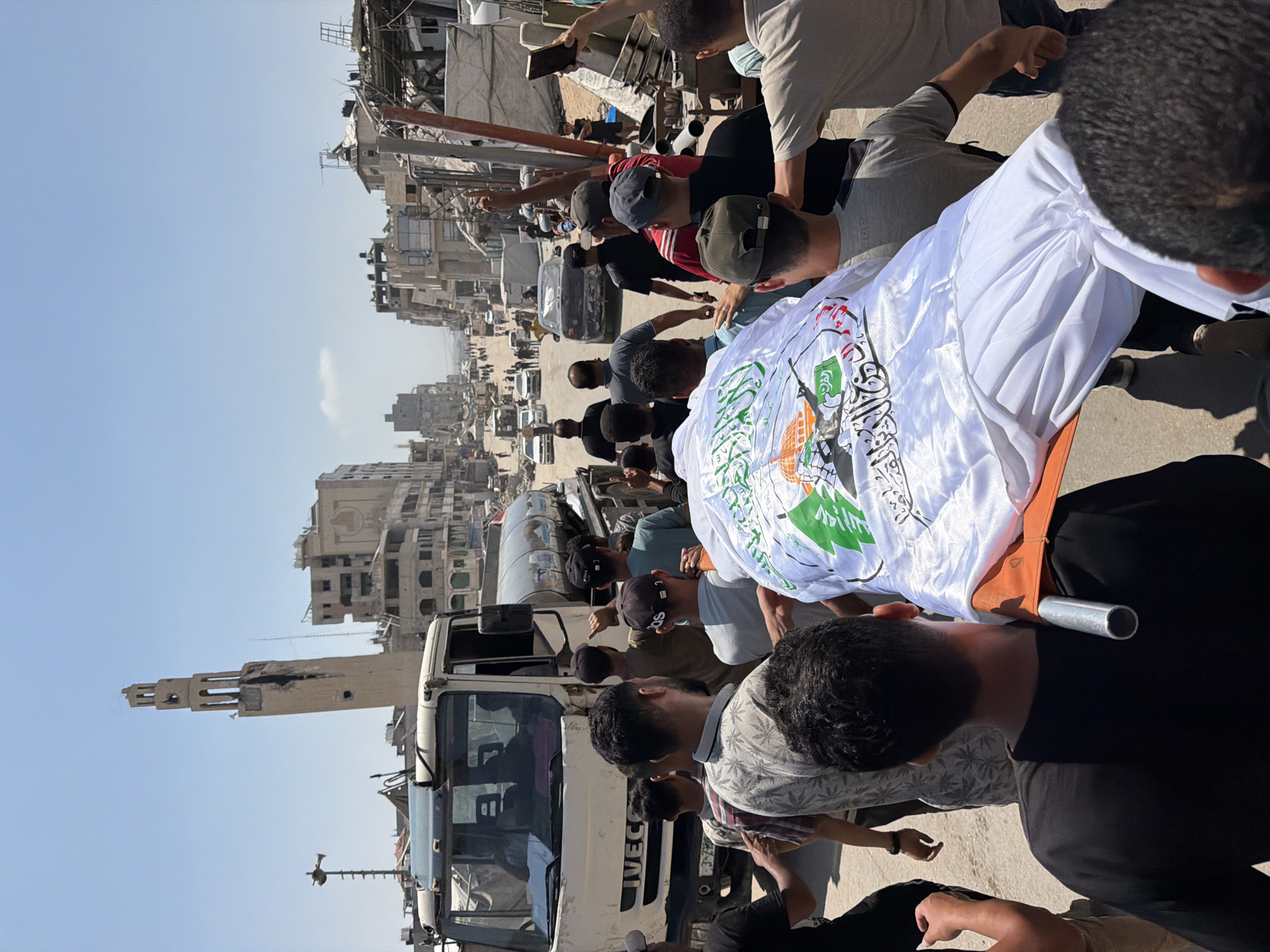 A group of men carry a deceased person on a stretcher through a street in the city of Gaza, Palestinian Territories. The deceased is covered in a white shroud adorned with an image of a mosque and Arabic text. The surrounding environment shows significant destruction, with damaged buildings and rubble lining the street. A white truck with a water tank is visible on the left side of the image, and several cars are present in the background. The sky is clear and blue.