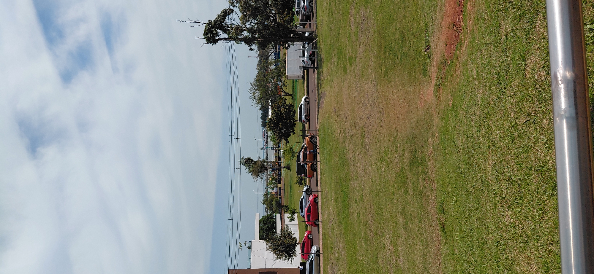 The image depicts a street scene in Encarnacion, Paraguay, with a focus on vehicles and a grassy area. In the foreground, a metal railing is visible, with a large expanse of green grass extending into the middle ground. Several cars are parked or driving along a paved road in the middle ground, including a red hatchback, a silver sedan, an orange pickup truck, and a white SUV. A person is walking near the orange pickup truck. In the background, there are trees, buildings, power lines, and a body of water with a ship visible on it. The sky is partly cloudy with a mix of white clouds and blue sky. The overall impression is of a typical urban street with a park-like area.