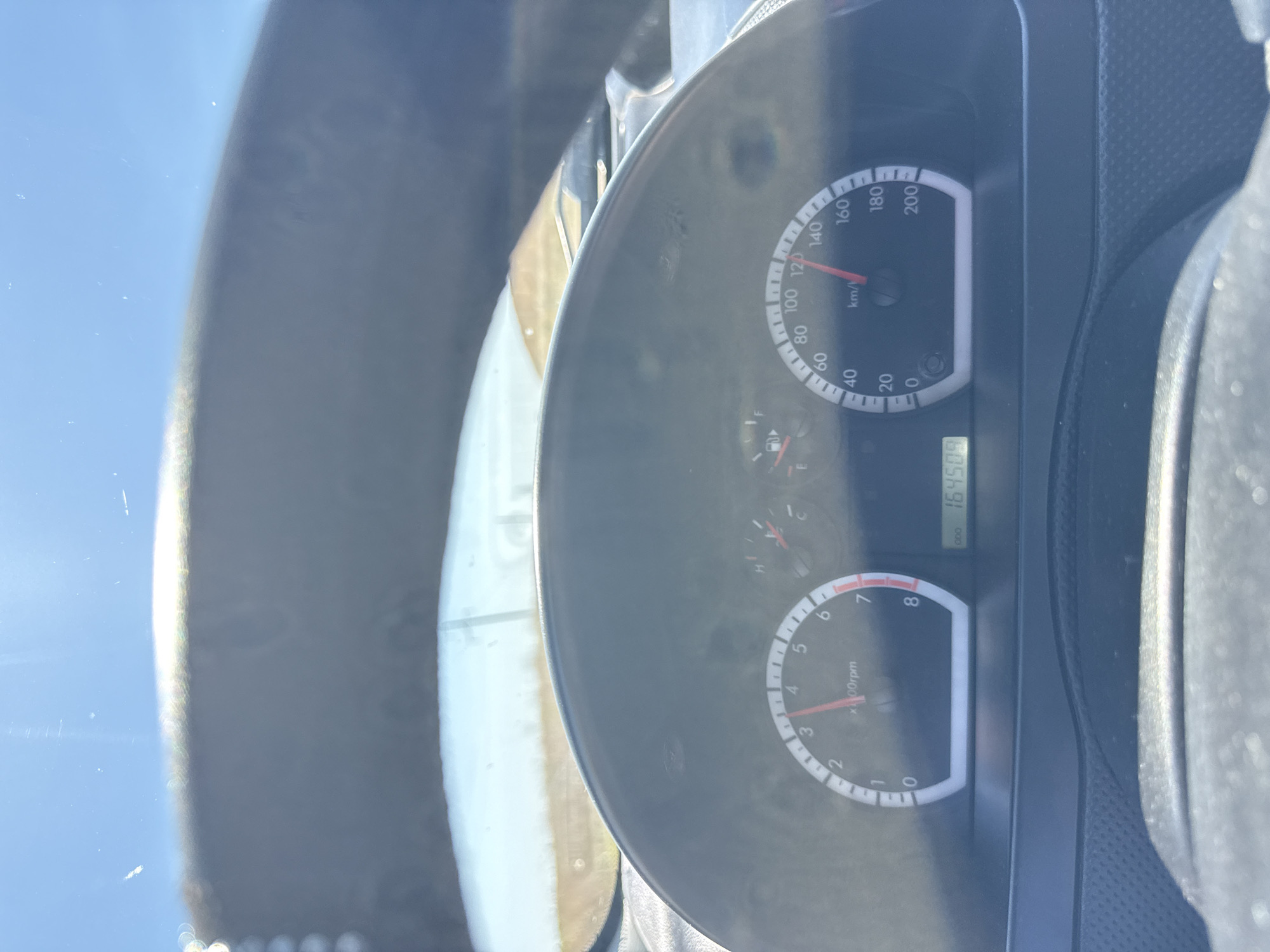 The image is a close-up, eye-level shot of a car's dashboard, taken from the driver's perspective. The dashboard displays a speedometer and a tachometer. The speedometer needle is pointing at approximately 115 km/h, and the tachometer needle is pointing at about 2.5 x 100 rpm. An odometer displays the mileage as 164509. The scene is illuminated by bright sunlight, suggesting it is daytime. The background shows a blurred view through the windshield of a bright sky and a hint of land. The specific location within the city of Cioc-Maidan, Moldova, is not visually discernible from the image itself.