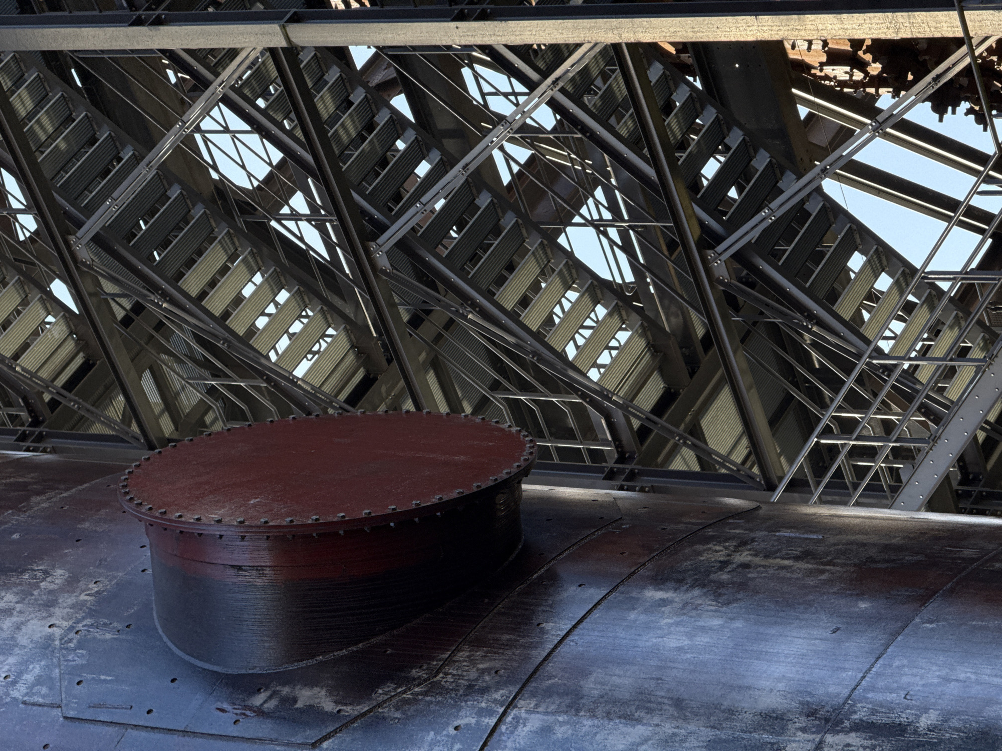 The image presents a detailed, low-angle view of industrial architecture, situated at the Belval heritage site in Esch-sur-Alzette, Luxembourg. On the left, a large, weathered, cylindrical metal structure, consistent with a blast furnace component, occupies the foreground. Its surface displays a dark blue-grey hue with extensive reddish-brown rust and discoloration, accented by vertical seams secured with bolts or rivets. A prominent, circular, dark reddish-brown metal port or capped pipe, secured by a series of visible bolts around its perimeter, projects horizontally from the cylinder's side. To the right and extending into the background, a complex, multi-level system of modern steel stairs, platforms, and structural beams is visible. The stairs feature open-grate or corrugated metal treads and are supported by a diagonal lattice of dark grey steel girders and uprights. Sections of a clear blue sky are visible through the gaps within the metal framework of the staircase. No people or discernible actions are depicted in the scene.