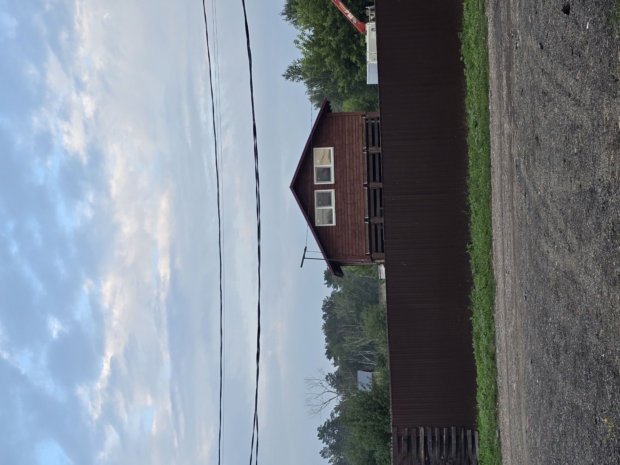 The image depicts a rural scene with a brown corrugated metal fence in the foreground. Behind the fence, a wooden house with a red roof is elevated on stilts, featuring two windows on its upper level. Further in the background, a dense forest of green trees stands beneath a cloudy sky. Overhead, two dark power lines stretch across the frame. A small section of a construction crane is visible to the right of the house. The ground in the foreground is covered with gravel. The scene appears to be located in Ермолаево, Russia.