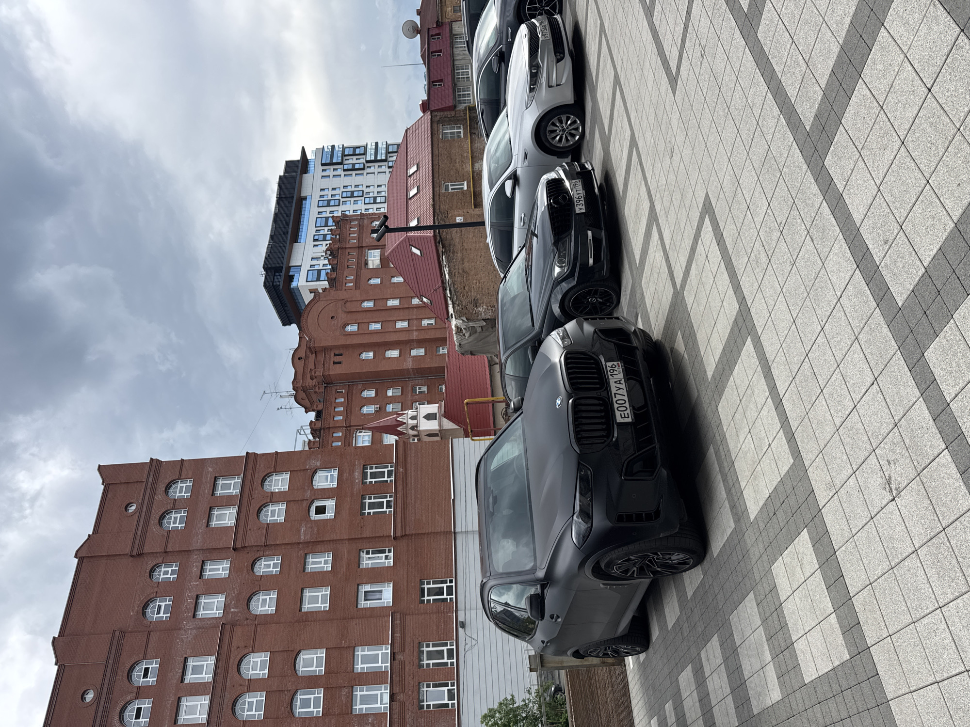 The image displays a line of parked cars on a tiled walkway in front of several buildings. The cars are oriented diagonally across the frame, with the closest car being a dark-colored SUV. Its license plate is visible and reads "E007YA 196". Behind it is a lighter-colored sedan, followed by another dark-colored sedan and a silver car.
The buildings in the background vary in architectural style and material. On the left, there is a tall, modern white and blue building. Next to it is a red brick building with a distinctive stepped roofline and numerous windows. Further back, another red brick building, possibly older, is visible. The sky is overcast, suggesting cloudy or potentially rainy weather, and the overall lighting indicates it is daytime. The tiled walkway is laid out in a geometric pattern of light and dark gray squares. The scene appears to be a street or parking area in an urban setting. No people are visible in the image.