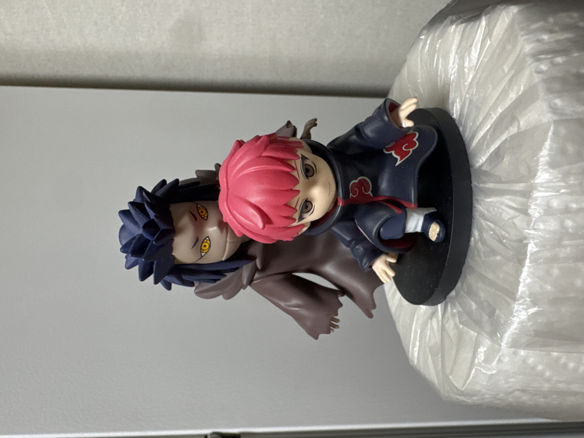This is a close-up, eye-level shot of two anime figurines, likely from the Naruto series, positioned on a black circular base. The figurine in the foreground has bright pink spiky hair, a dark blue cloak with red cloud emblems, and light skin. It is standing with its left leg slightly forward. Behind and slightly to the left of the foreground figurine is a second figurine with spiky dark blue hair and bright yellow eyes with slit pupils. This figurine is wearing a dark brown, flowing cloak. Both figurines are facing forward. The base is resting on a white, textured surface that appears to be a roll of plastic wrap or similar material. The background is a plain, light gray wall. The lighting is even and appears to be artificial. The image was captured indoors, likely in a residential setting. No specific landmarks or elements indicative of Anseong-si, South Korea are visible in the image.