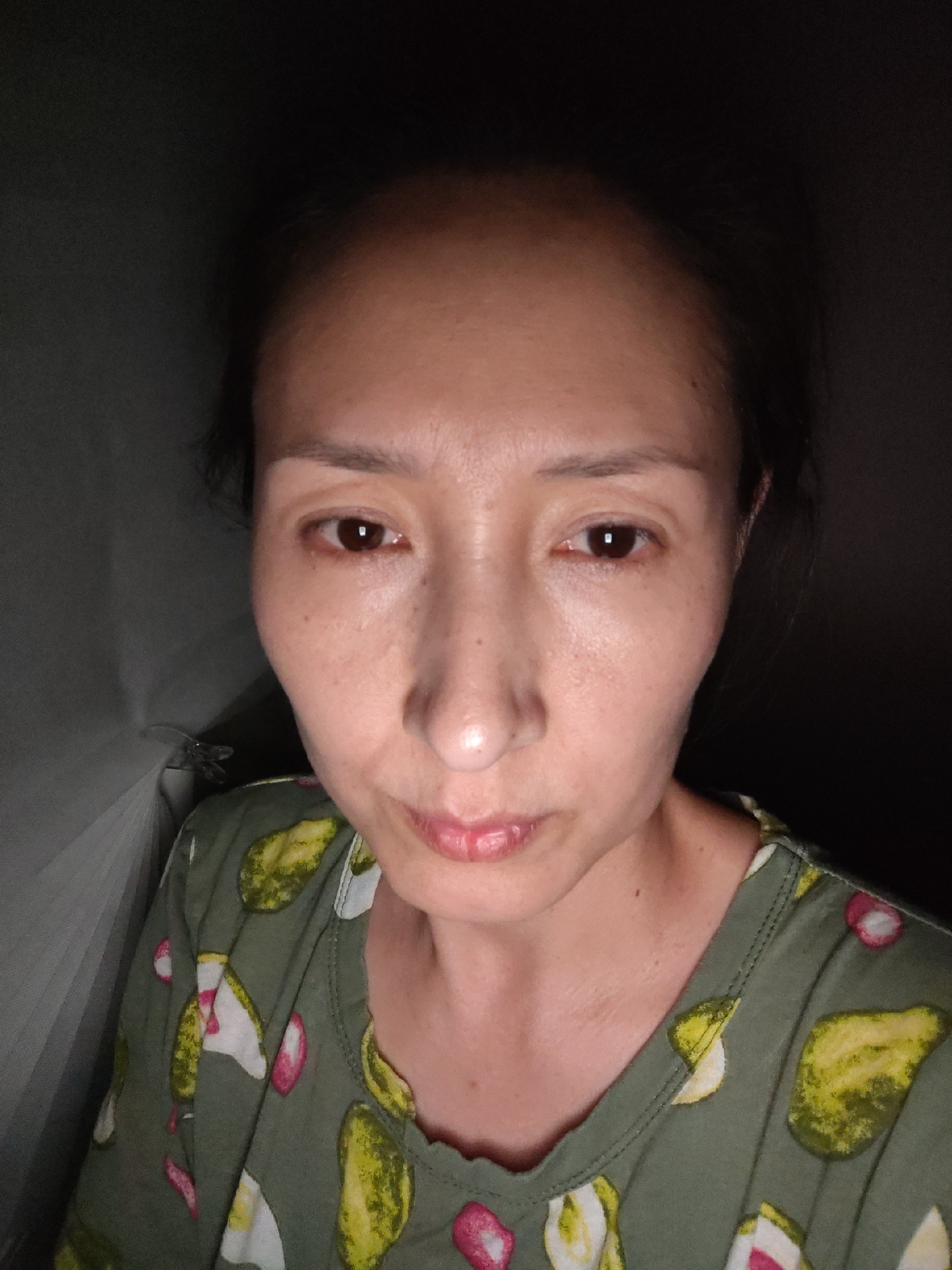The image is a close-up, head-and-shoulders shot of an East Asian woman. She is looking directly at the camera. Her skin is pale and appears to have some texture. Her hair is dark and pulled back. She is wearing a green shirt with a pattern of what appears to be pickles and pink and white shapes. The background is dark and appears to be indoors, possibly a room with a textured wall or curtain on the left. There is no indication of the city of Oral, Kazakhstan in the image.