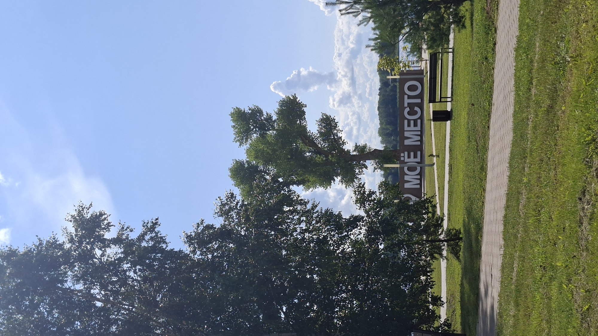 The image depicts a park scene on a sunny day in Myadzyel, Belarus. In the foreground, a paved walkway runs horizontally across the frame, bordered by well-maintained green grass. To the left and right of the walkway, more grassy areas are visible, with dappled sunlight indicating the presence of trees.
Dominating the middle ground is a large, three-dimensional sign with the Cyrillic text "МОЁ МЕСТО" (My Place). The letters are white and appear to be made of a solid material, possibly wood or metal, and are mounted on a dark brown background structure. To the right of the sign, a dark-colored park bench with a slatted seat and back is placed on the grass. Behind the sign and bench, a body of water, likely Lake Myadzyel, is visible in the distance, with a tree-lined shore forming the horizon.
Several trees are present in the scene. A large, leafy tree with dark green foliage stands prominently on the left side of the image, partially obscuring the sky. In the middle of the frame, a tall tree with a more open canopy and lighter green leaves stands behind the "МОЁ МЕСТО" sign. To the far right, a coniferous tree adds to the park's greenery.
The sky is a vibrant blue with scattered white, fluffy clouds. One notable cloud formation in the upper right portion of the sky has an unusual, almost anthropomorphic shape. The overall impression is one of a peaceful, well-kept public space, likely a recreational area near the lake. No people are visible in the image.