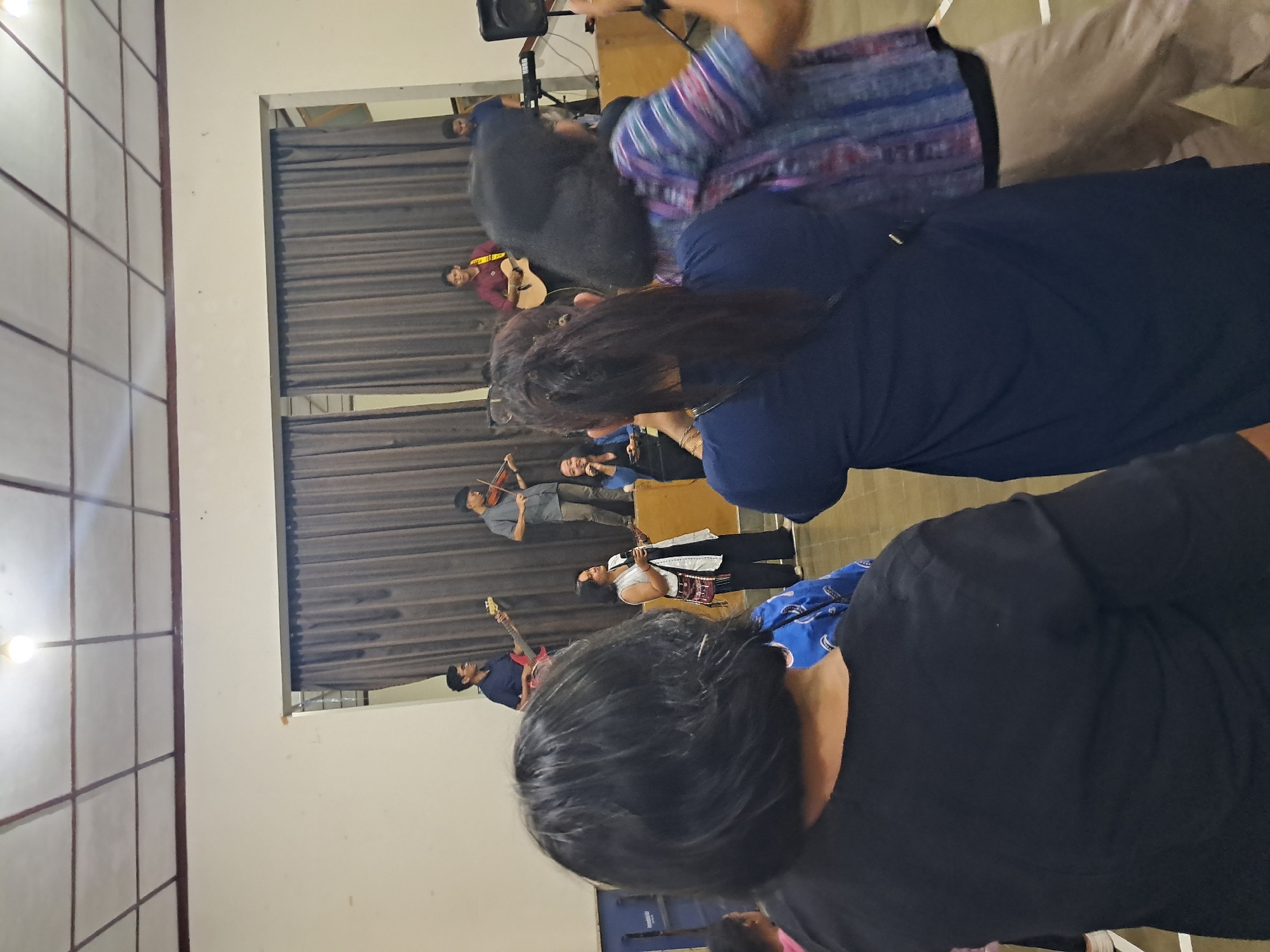 This image depicts an indoor gathering in Ngaglik, Indonesia, likely a community event or performance. In the background, on a raised stage area, a band is performing. A man in a blue shirt plays a red electric guitar, and another man in a grey shirt plays a violin. A woman in a black top and pants, wearing glasses, is also on stage, holding a microphone and appearing to sing or speak. To the right of the stage, a person in a maroon shirt plays an acoustic guitar, and another person in a blue shirt is also present. A keyboard and a speaker are visible on the stage. In the foreground, a diverse group of people are watching the performance. The focus is on the backs of several individuals, including a woman with short black hair in a black shirt, and another woman in a dark blue shirt. To their right, a person in a colorful, patterned shirt and tan pants is also visible. The environment appears to be a large room with a light-colored ceiling and brown curtains behind the stage. The overall atmosphere suggests a casual and engaging event.