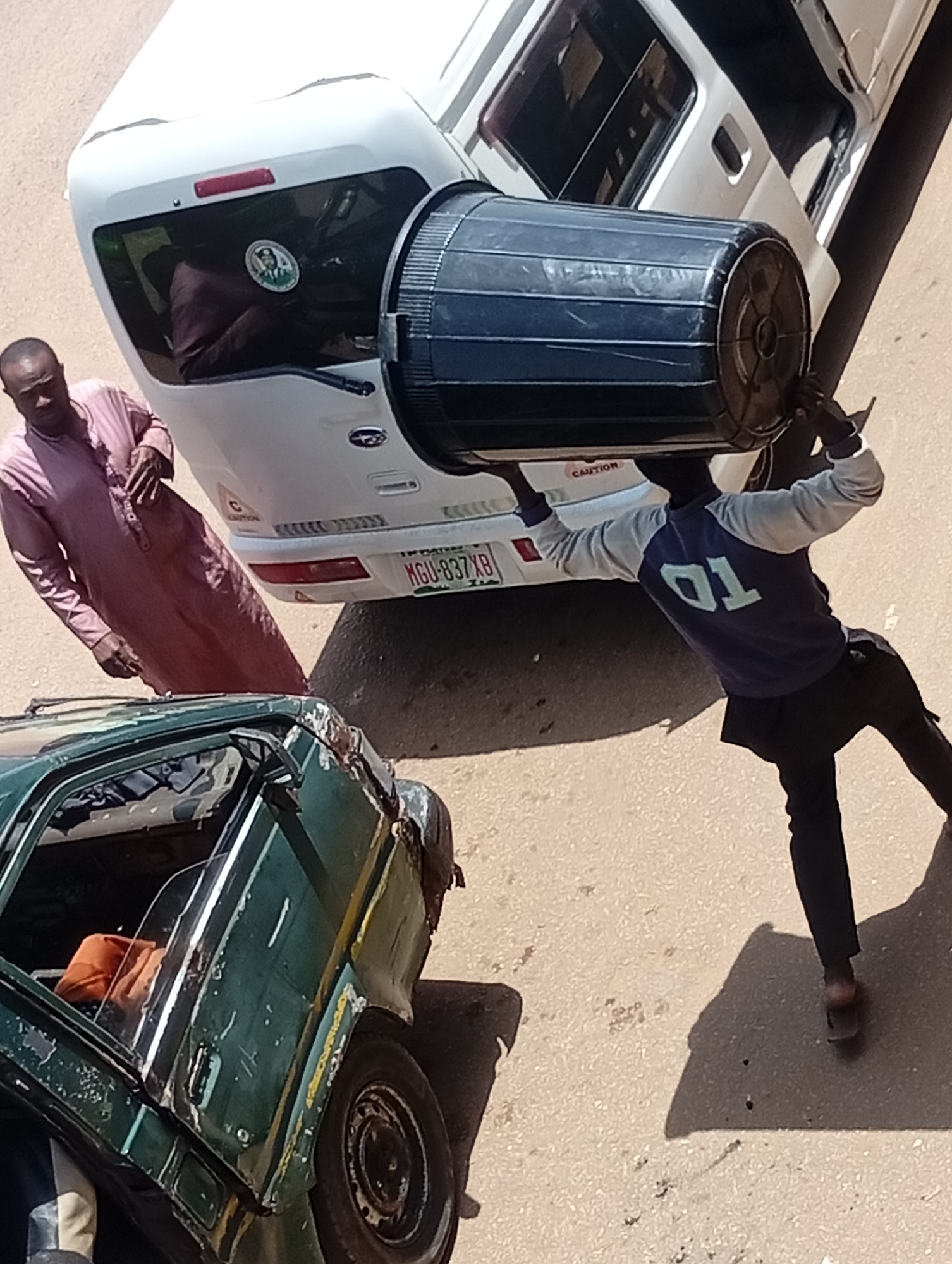 In Bukuru, Nigeria, a man in a pink thobe stands near the rear of a white van, looking towards the camera. A second man, wearing a blue and gray long-sleeved shirt with "01" on the back and dark pants, is positioned under the van, holding a large black cylindrical object. He appears to be lifting or maneuvering the object from beneath the vehicle. In the foreground, a green, heavily damaged car with its door open reveals an orange fabric item inside. The scene takes place on an asphalt surface, likely a street or parking area, with shadows cast by the vehicles and people.
