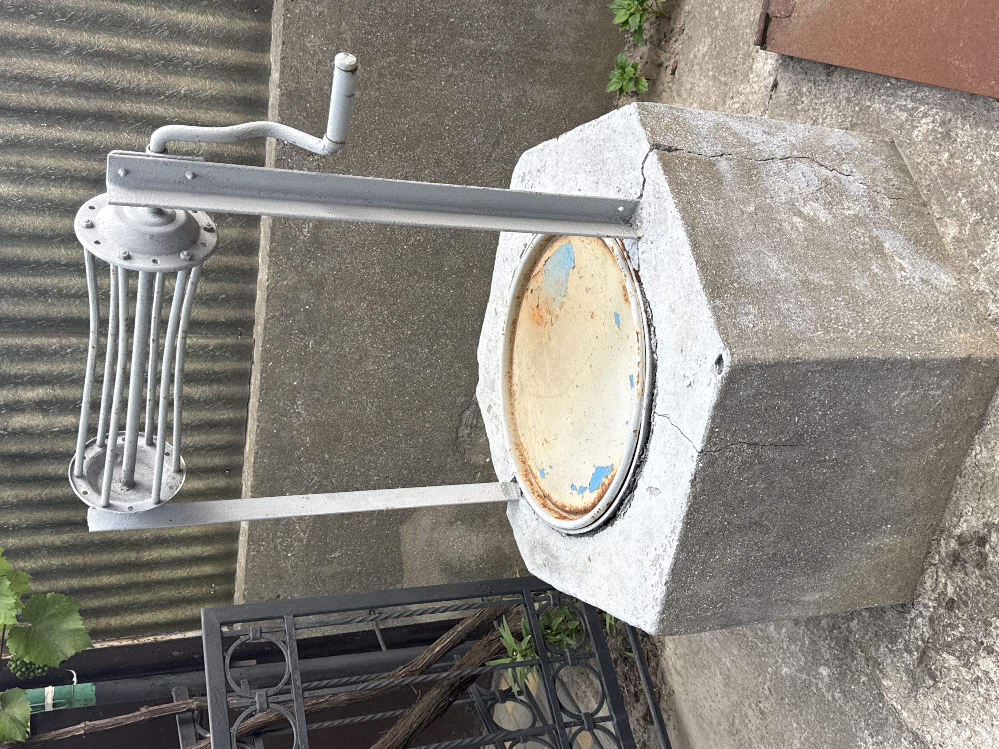 An outdoor, eye-level, full shot shows a concrete well with a metal crank and reel assembly. The well's base is a hexagonal concrete structure with visible cracks and chips. A weathered, circular metal cover rests on top of the well, exhibiting rust and peeling blue paint. The reel assembly, painted light gray, has a crank on the right side and several horizontal bars. Behind the well, a corrugated metal fence forms the background, with a concrete ledge visible at its base. To the left, a black metal fence and a dark pole with green leaves and grapes are partially visible. Green plants grow at the base of the well and to the right.