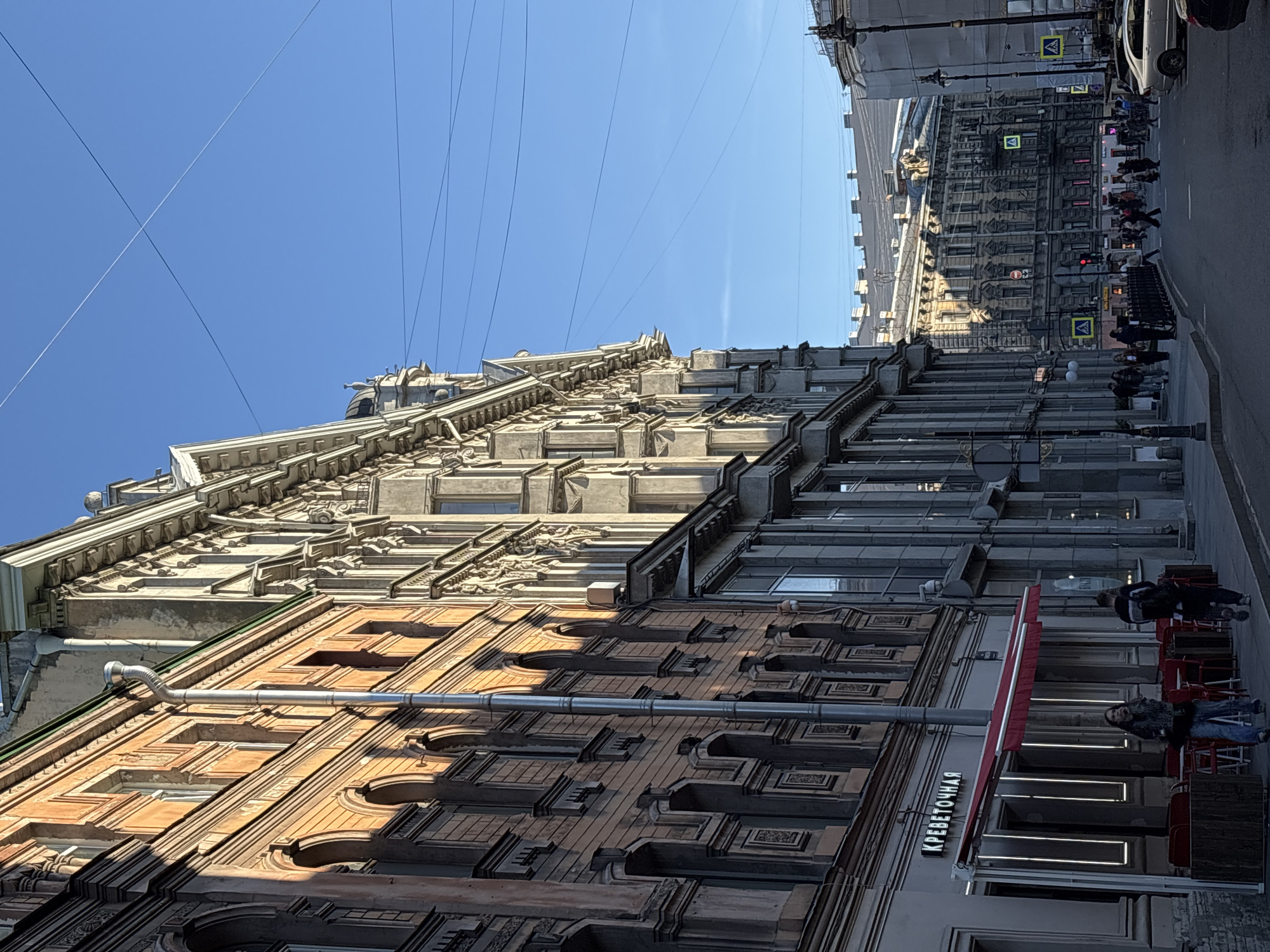 The image depicts a street scene in Saint Petersburg, Russia. Several buildings of varying architectural styles are visible. One building on the left has an ornate facade with detailed carvings and an orange-brown exterior. Another building to the right is taller and features a more streamlined design with numerous windows and grey tones.
Wires stretch across the clear blue sky above the buildings.
On the right side of the image, the street is visible with pedestrians walking, traffic lights, street lamps and crosswalk signs. A sidewalk café with red awning is visible, some people are sitting at red chairs. The sign shows "КРЕВЕТОЧНАЯ" in Cyrillic script.
The image orientation is slightly tilted.