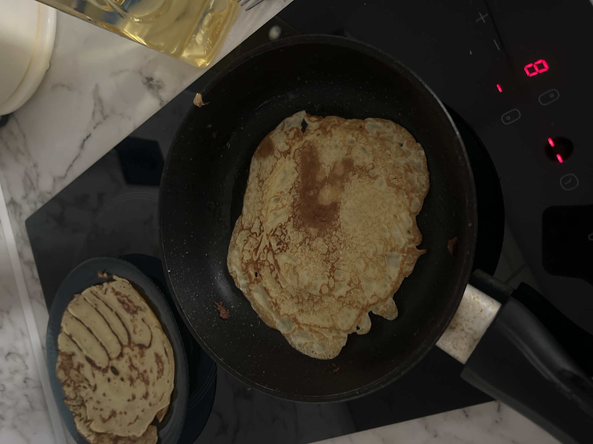A close-up, overhead shot shows crepes being cooked in a pan on a stovetop. A plate of already-cooked crepes is visible to the left, and a bottle of oil is on the counter to the right. The stovetop has digital controls, with a red "8" displayed on one of the burners. The scene appears to be in a kitchen, likely in Krasnodar, Russia, based on the implied location. The lighting is dim, with a focus on the food and cooking apparatus. There are no people or animals present in the image.