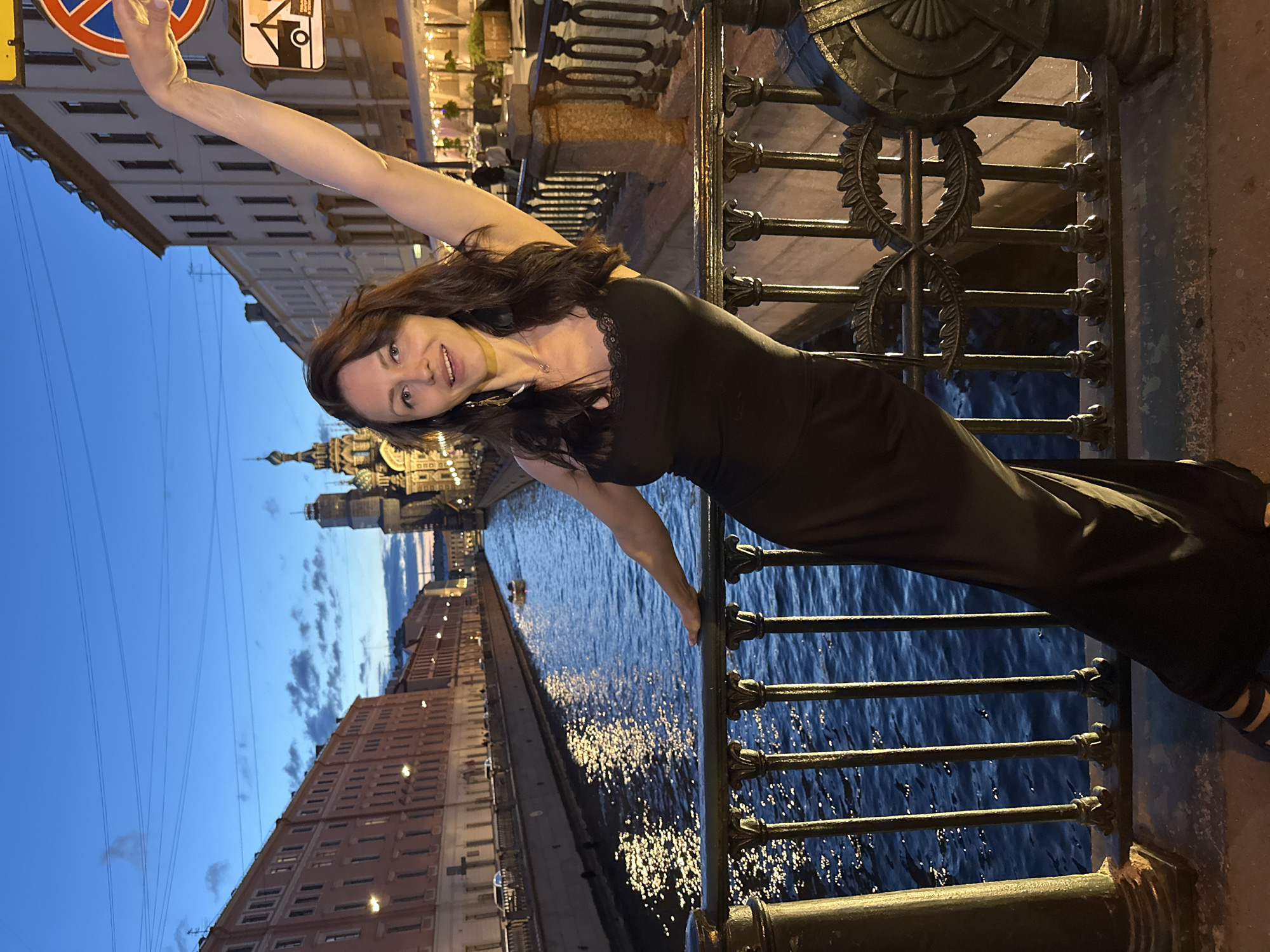 The image captures a woman standing on a bridge at dusk in Saint Petersburg, Russia. She is wearing a black dress and has her right arm raised. Behind her, a canal is visible, reflecting the lights of the buildings lining its banks. The Church of the Savior on Spilled Blood, a prominent landmark, can be seen in the background, its domes illuminated against the twilight sky. The scene appears to be along the Griboyedov Canal.