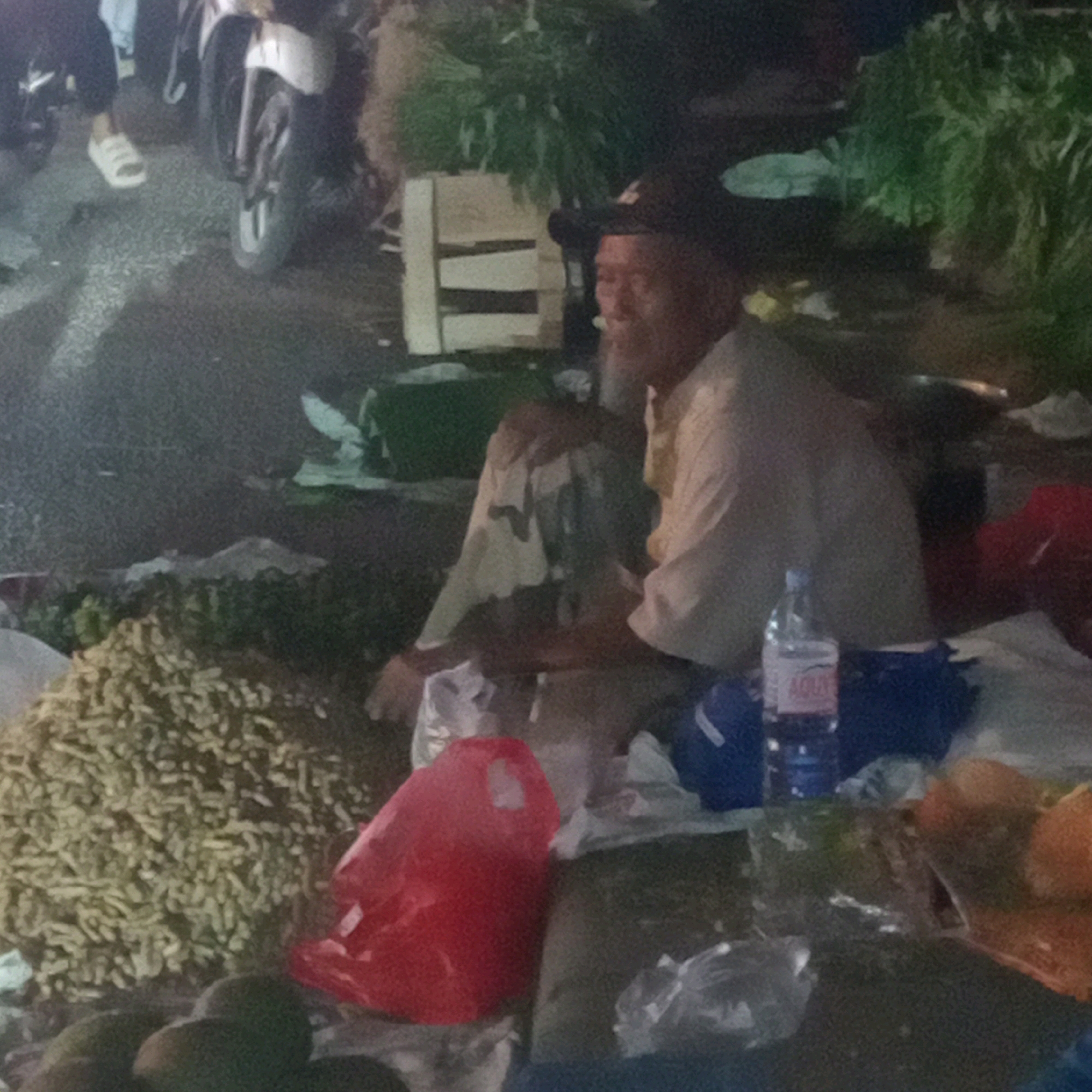 The image captures a scene at night, possibly a roadside market or gathering, in Cileungsi, Indonesia. In the foreground, a man wearing a dark hat and a light-colored shirt is seated. His face is visible from the side, and he has a beard. To his left, a large pile of small, light-brown items, possibly nuts or seeds, is spread out on a dark surface. To the right of the man, a clear plastic bottle with a red label is visible, resting on a dark blue object. Further to the right, there are some orange-colored fruits, possibly tangerines or oranges, and a red plastic bag. In the background, a motorcycle with a white front fender is partially visible, along with other motorcycles and people dressed in dark clothing. There are also some green plants or foliage in the background, suggesting a possible outdoor setting. The ground appears to be paved, and the overall lighting suggests it is nighttime, with artificial illumination casting shadows.