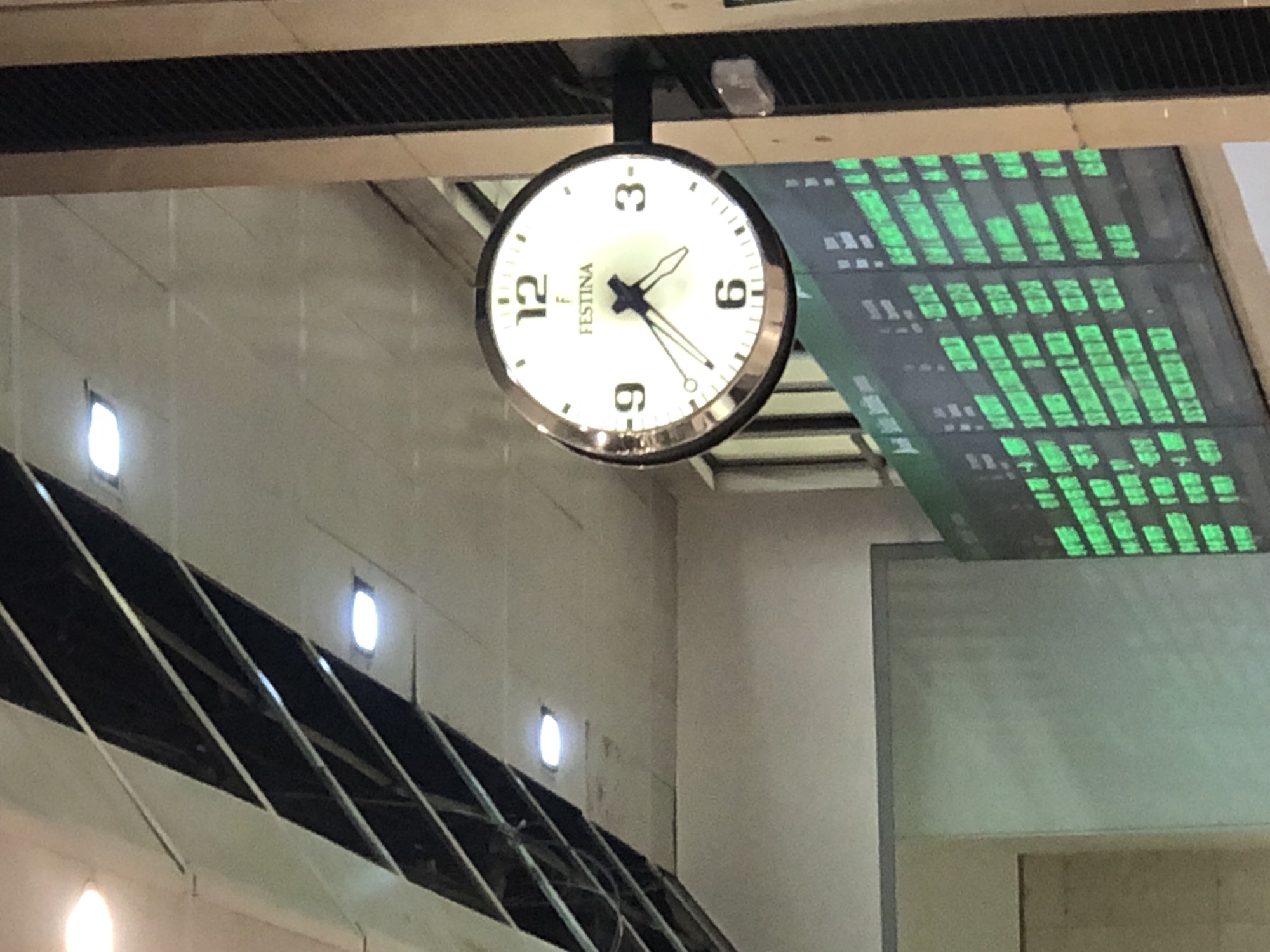 The image shows a round Festina clock with a white face, black numerals, and black hands, mounted on a vertical pole. The clock is illuminated from behind. To the right of the clock, there is a digital display board with green text. The background consists of a ceiling with diagonal beams and recessed lighting. The scene appears to be an indoor public space, possibly a train station or airport. The time on the clock appears to be approximately 10:47.
