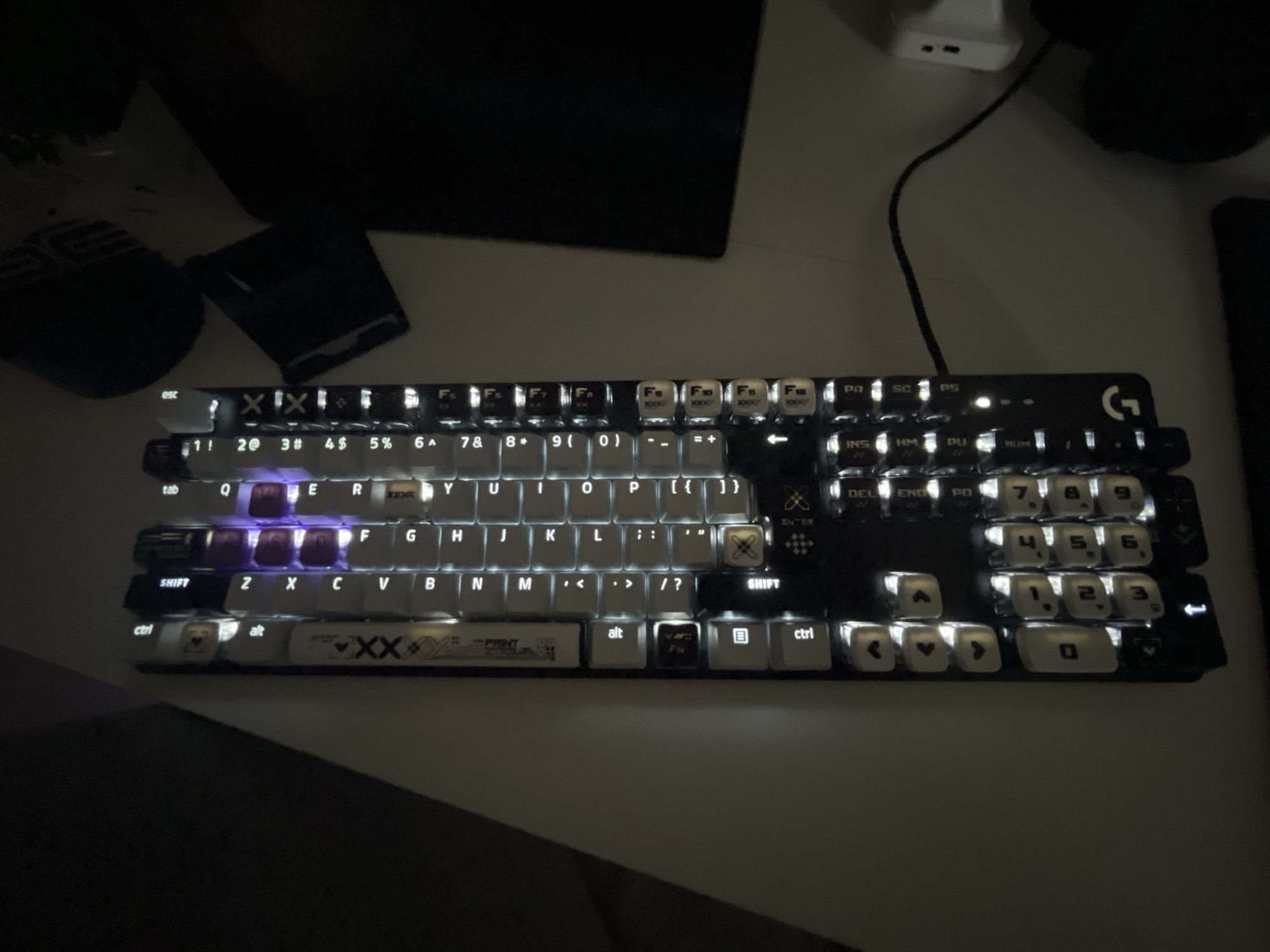 The image shows a close-up of a black and white mechanical keyboard with white backlit keys, illuminated in a dimly lit room. The keyboard is positioned on a white surface, likely a desk. A dark object, possibly a monitor or laptop, is visible in the background, casting shadows. A white power adapter with two USB ports and a black wire are also present, suggesting an electronic setup. The lighting is low, creating a moody atmosphere. The keyboard itself is the primary focus, showcasing its layout and illuminated keys. Some keys display symbols and letters, and there's a Logitech logo on the top right. A prominent graphic of two intertwined "X" symbols is visible on the spacebar and several other keys. The overall scene suggests a personal computer setup, likely used for gaming or work, in a domestic or office environment. The setting is a desk in Varna, Bulgaria. There are no people visible in the image.