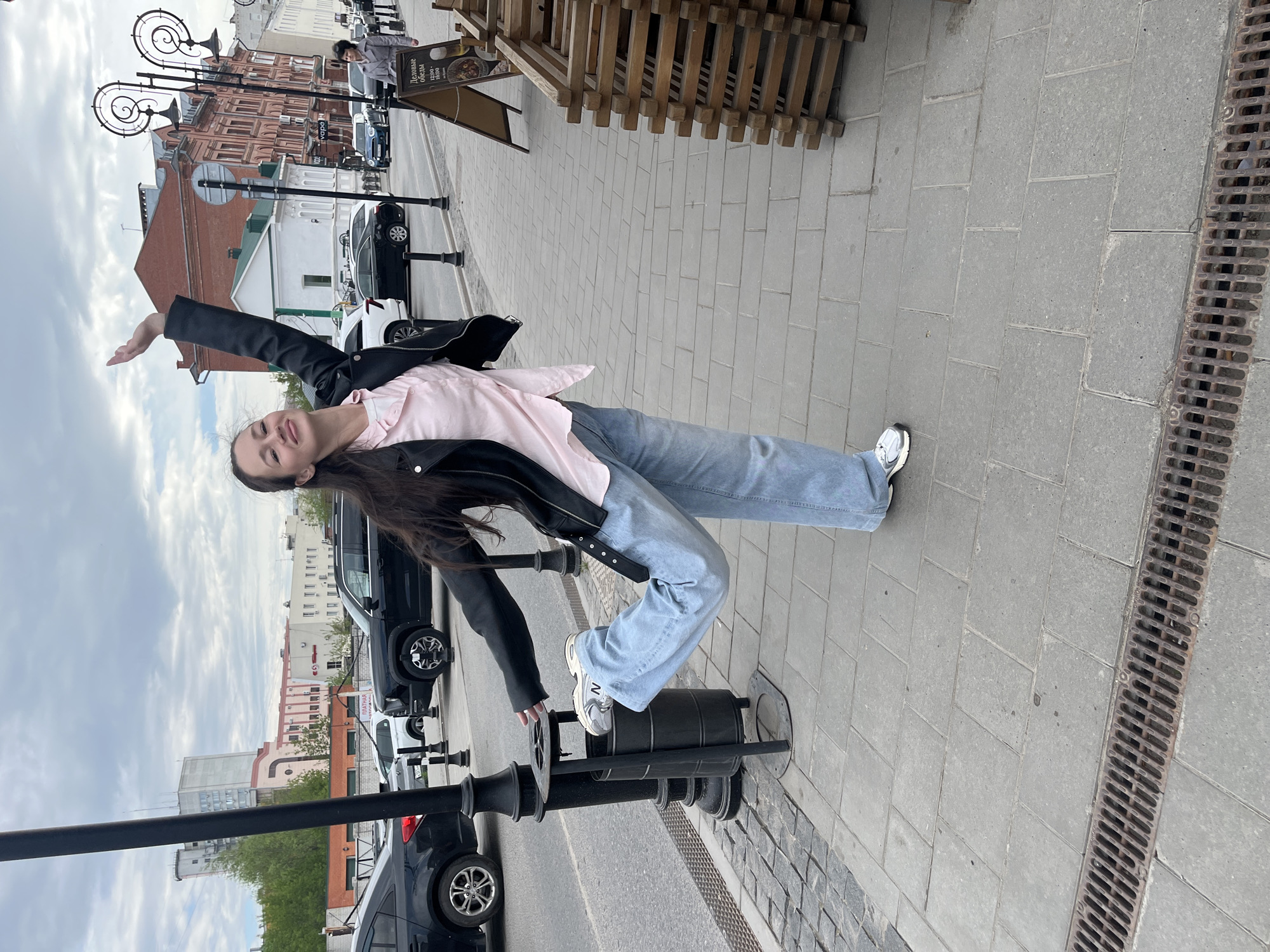 A young woman poses on a sidewalk in Ulyanovsk, Russia. She is leaning back, supported by a black metal bollard. She is wearing a pink shirt, light blue jeans, a black leather jacket, and white sneakers. She has long brown hair and is smiling. Cars and buildings are visible in the background. A metal storm drain is near the edge of the sidewalk.