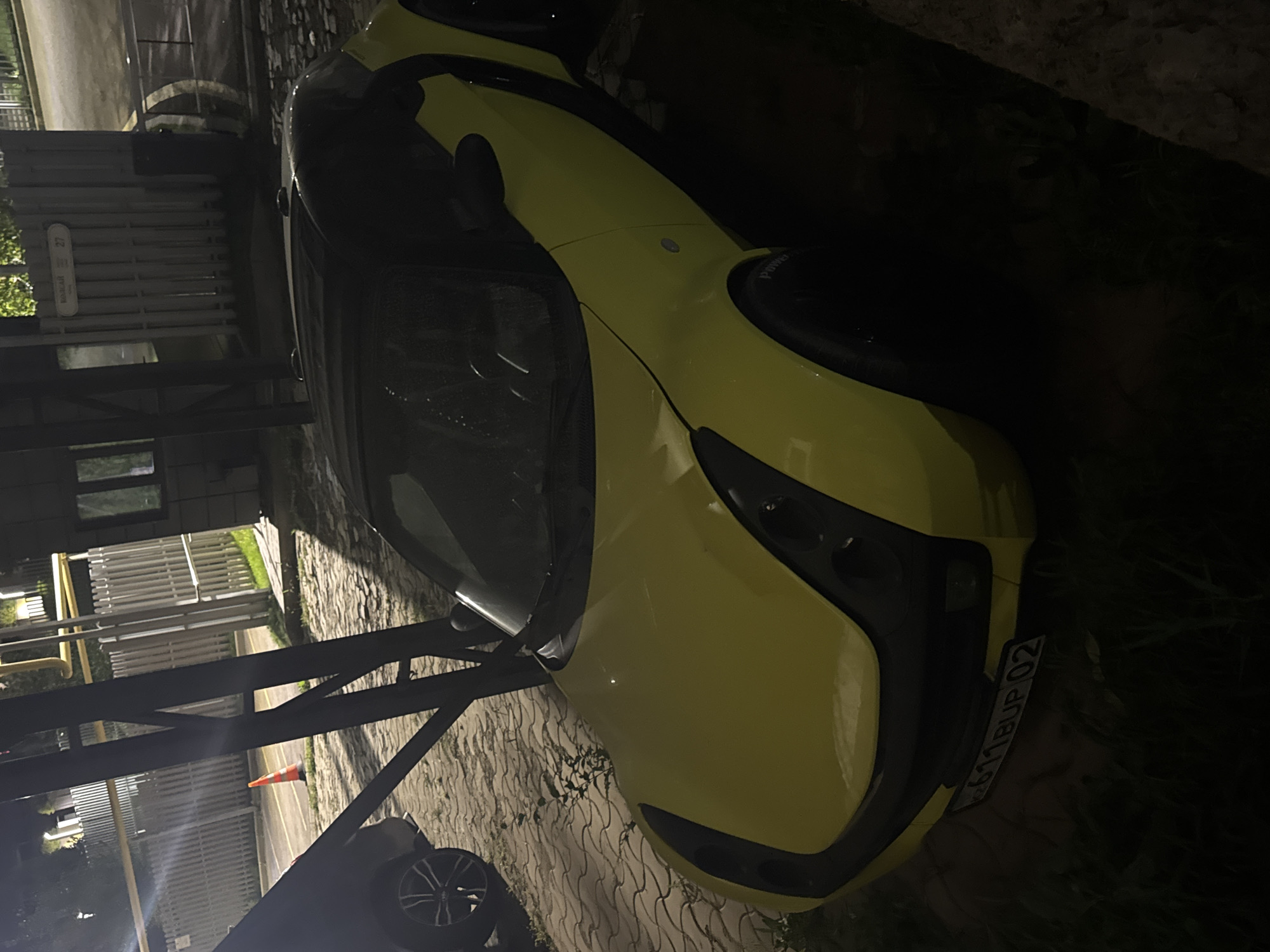 The image shows a yellow Smart car parked on a paved surface, possibly a sidewalk or parking area, with grass and foliage visible to the sides. The car has a black soft top and black accents around the headlights and wheel wells. A license plate with Cyrillic characters and numbers is visible on the front of the car. In the background, there is a metal fence and a building with a sign that reads "BONCAR 27". There is also an orange traffic cone visible in the distance near the fence. The scene appears to be outdoors at night or in low-light conditions, with artificial lighting illuminating parts of the area. The location is identified as Almaty, Kazakhstan.