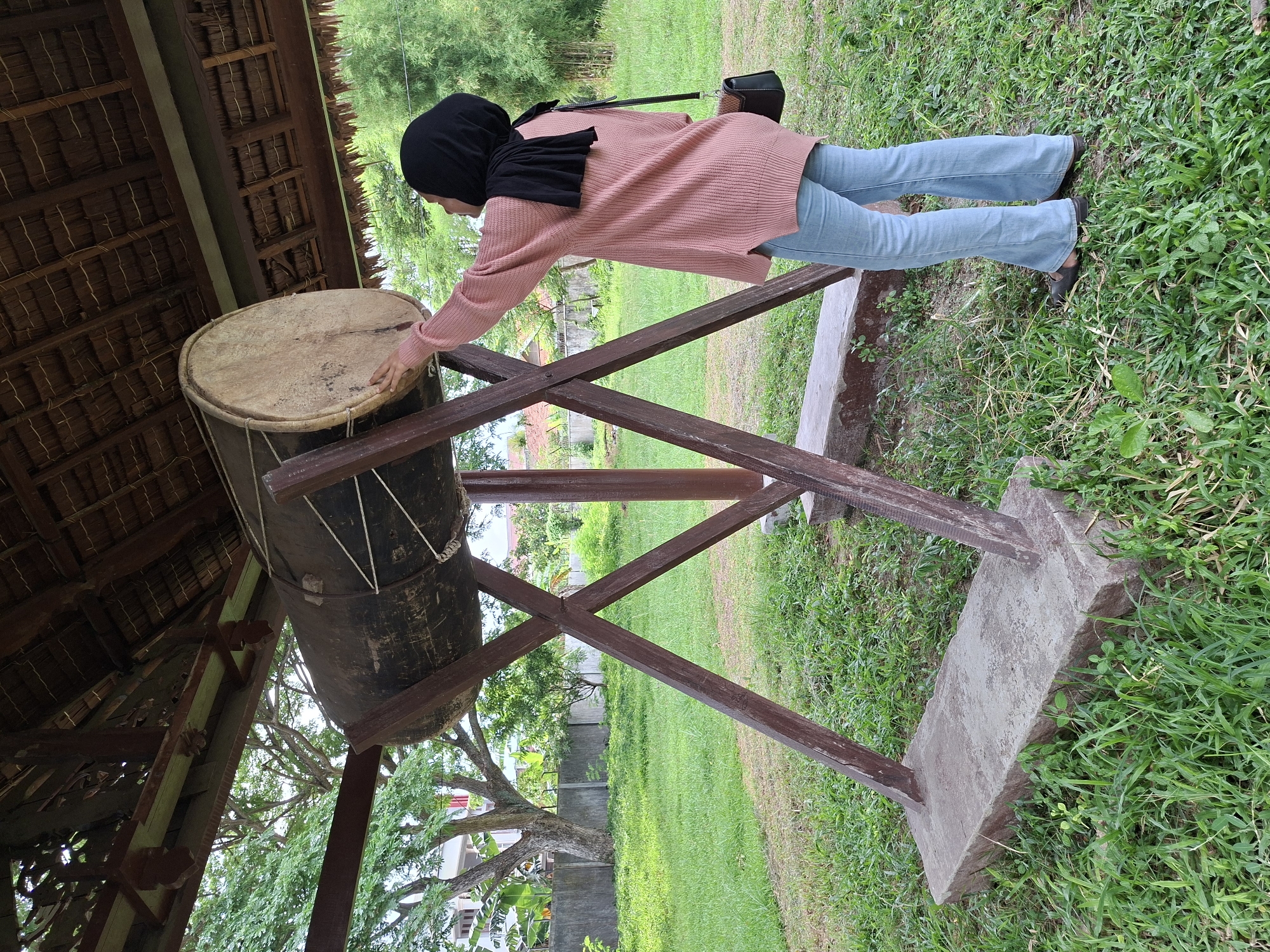 A woman in a pink sweater, blue jeans, and a black hijab stands touching a large drum supported by a brown wooden frame. The drum is cylindrical, dark brown on the sides, and has a lighter, circular top. The structure holding the drum is placed on a concrete base, surrounded by green grass. The scene is partially shaded by a roof structure with a thatched ceiling. The backdrop includes trees and other greenery, suggesting a park or outdoor space. The location is identifiable as Lhokseumawe, Indonesia, based on the architectural style and typical Southeast Asian environment.
