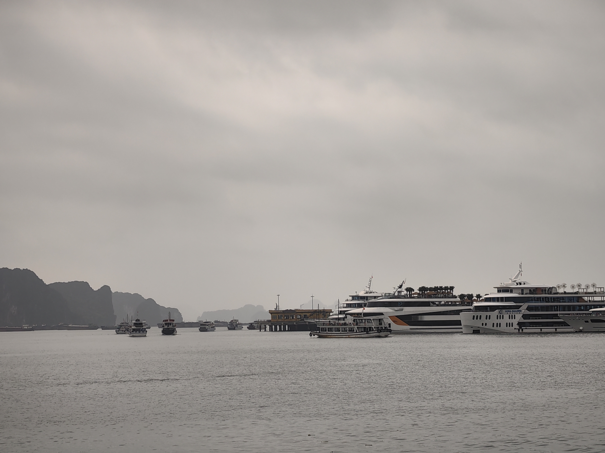 The image shows a seascape view in Bai Chay Ward, Vietnam, under a cloudy sky. The water is calm with slight ripples. Several boats are present. In the foreground, there are two large, multi-decked cruise ships docked, one painted with orange and white stripes, the other primarily white. A smaller tour boat is positioned closer to the viewer. In the mid-ground, a dock structure is visible with smaller vessels nearby. Further back, several smaller boats are scattered across the water. Distant limestone karsts formations characteristic of Ha Long Bay can be seen on the horizon. The overall color palette is muted due to the overcast weather. No people are distinctly visible in the image.