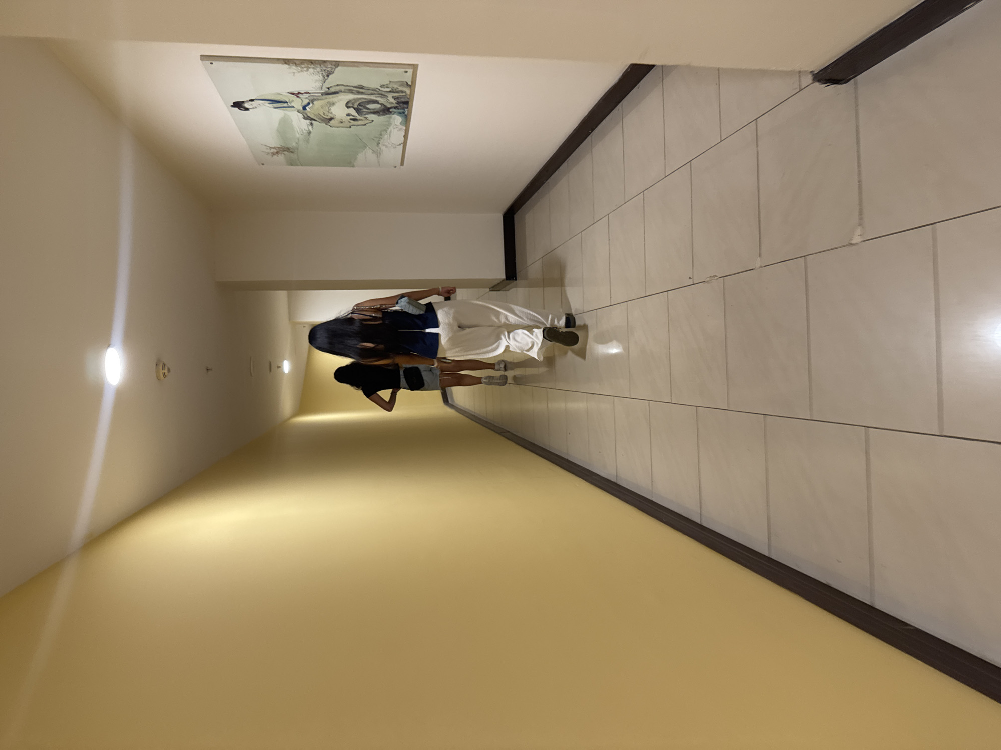 In a brightly lit hotel corridor in Aksu, Turkey, two women walk away from the camera. The corridor is long and narrow with pale yellow walls on the left and beige walls on the right. The floor is tiled with large, square, light-colored tiles. Recessed lighting fixtures are visible in the ceiling, casting a warm glow. On the right wall, a framed artwork depicting a woman in traditional attire standing on a rock with a misty background is hung. The woman in the foreground wears white linen pants and a dark blue sleeveless top, carrying a light blue purse. The second woman, further down the corridor, wears a black t-shirt and a light-colored denim skirt, with a black fanny pack around her waist. Both women have dark hair. The overall impression is one of quiet movement within a well-maintained, modern interior space.