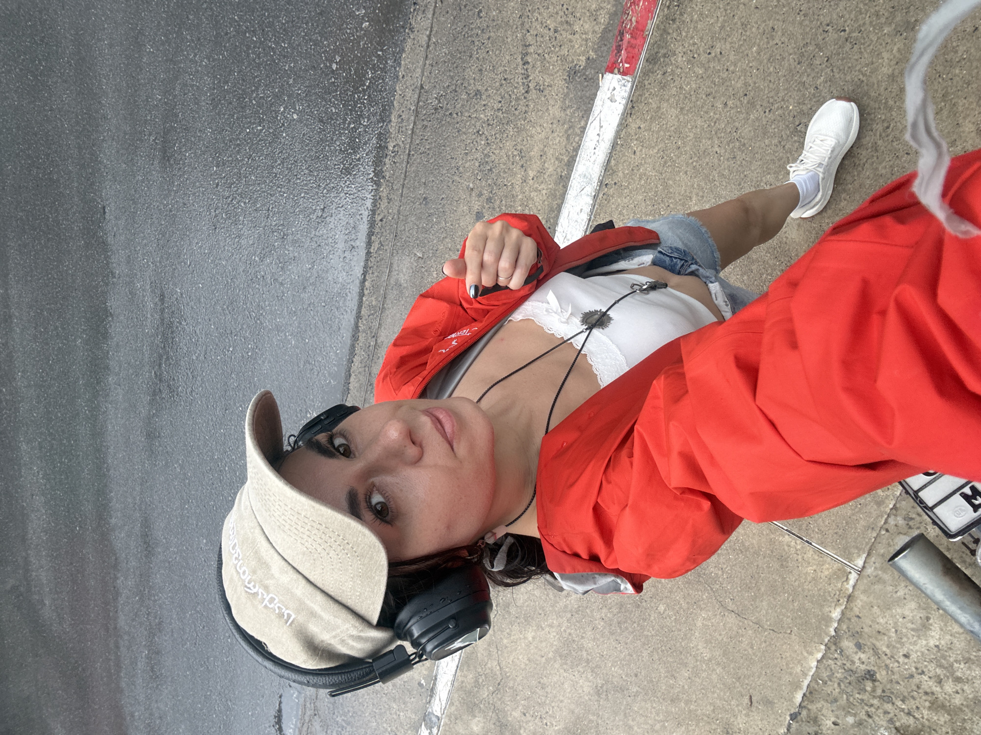 A woman wearing a beige baseball cap, black headphones, a white lace-trimmed top, denim shorts, a red rain jacket, and white sneakers is taking a selfie. She is standing on a wet, asphalt road in Karon, Thailand, near a curb with red and white markings. The weather appears to be rainy. She has her right hand raised, pointing her thumb upwards in a gesture.