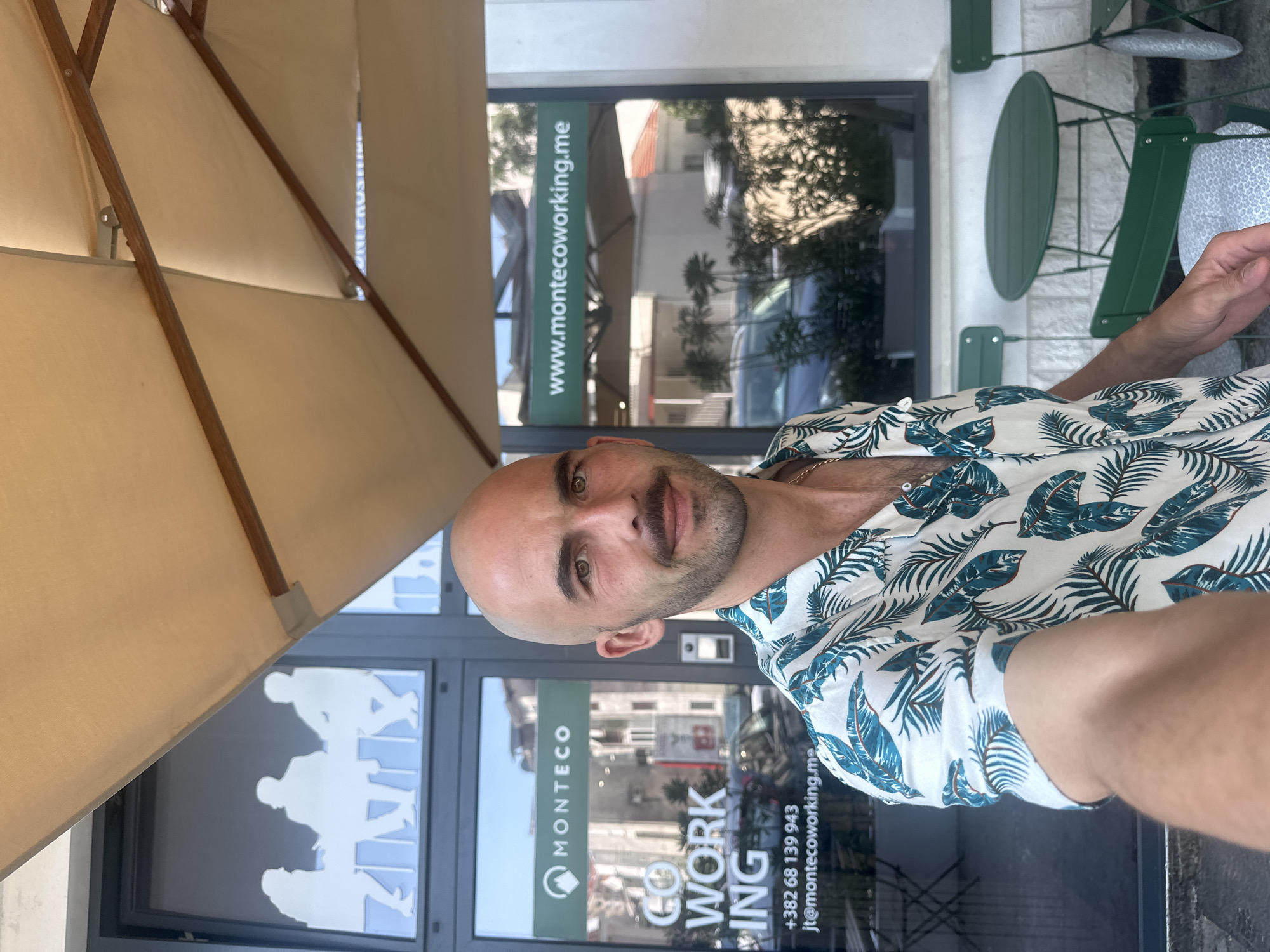 A man is positioned in front of a business identified as "MONTECO Coworking" in Budva, Montenegro. The business's contact information, including website and phone number, is visible on the storefront. The man is under an umbrella and near an outdoor green table and chair. He is wearing a light-colored shirt with a leaf pattern.