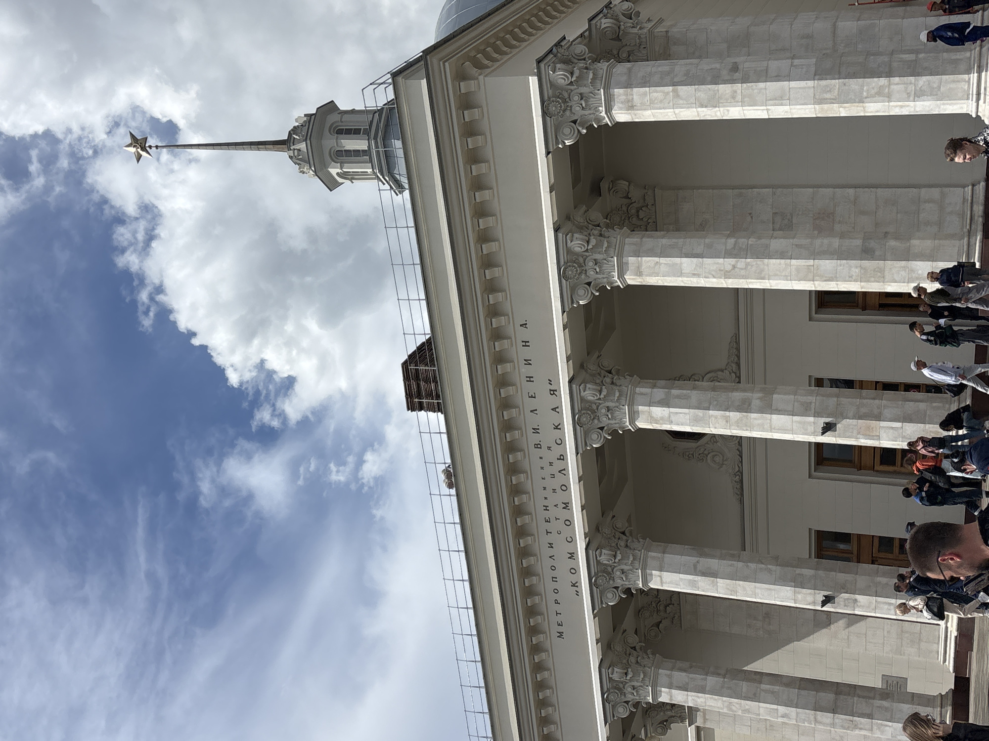 The image shows a building in Moscow, Russia. Specifically, it is the Komsomolskaya-Koltsevaya station of the Moscow Metro. The photograph is taken from below, looking up at the building's facade and spire. The facade has tall, classical columns. Inscribed on the facade are the words "METROPOLITEN IM. V. I. LENINA, STANTSIIA, 'KOMSOMOLSKAYA'." A spire topped with a five-pointed star rises into the sky which is filled with clouds. People are visible at the base of the building.