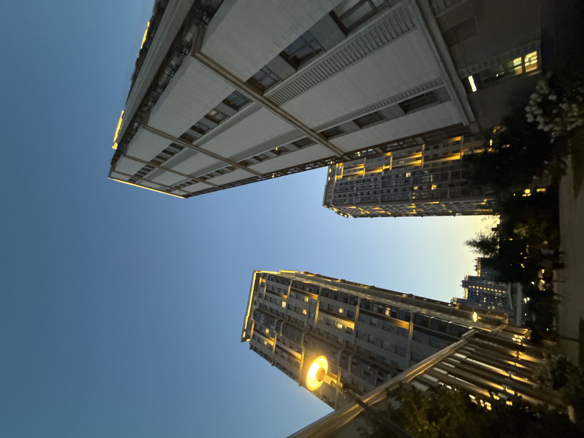 The image captures an urban scene at dusk, likely in Moscow, Russia. Tall, modern residential buildings dominate the frame, viewed from a low angle, creating a sense of their imposing height against a clear, pale blue sky. The buildings are illuminated with warm, artificial lights from windows and along their upper edges, suggesting that evening is settling in.
One of the prominent structures, a lighter colored building with a grid-like facade, is angled dramatically across the upper part of the image. Below it and to the right, another tall building, with a more uniform facade and rows of lit windows, stretches upwards. In the foreground, the top of a structure with angled metal beams is visible, likely part of a balcony or walkway, also featuring warm lighting.
A streetlamp with a bright, yellow-orange glow is visible, casting light onto the buildings and foreground elements, further emphasizing the transition from day to night. Trees and some foliage are visible on the right side of the image, partially obscuring the view of the buildings and adding a touch of nature to the otherwise architectural landscape. The overall mood is serene and urban, with the warm lights providing a sense of life and activity within the buildings. There is no visible text in the image. The time of day is clearly dusk or early evening, with the sky indicating a clear day with no precipitation.