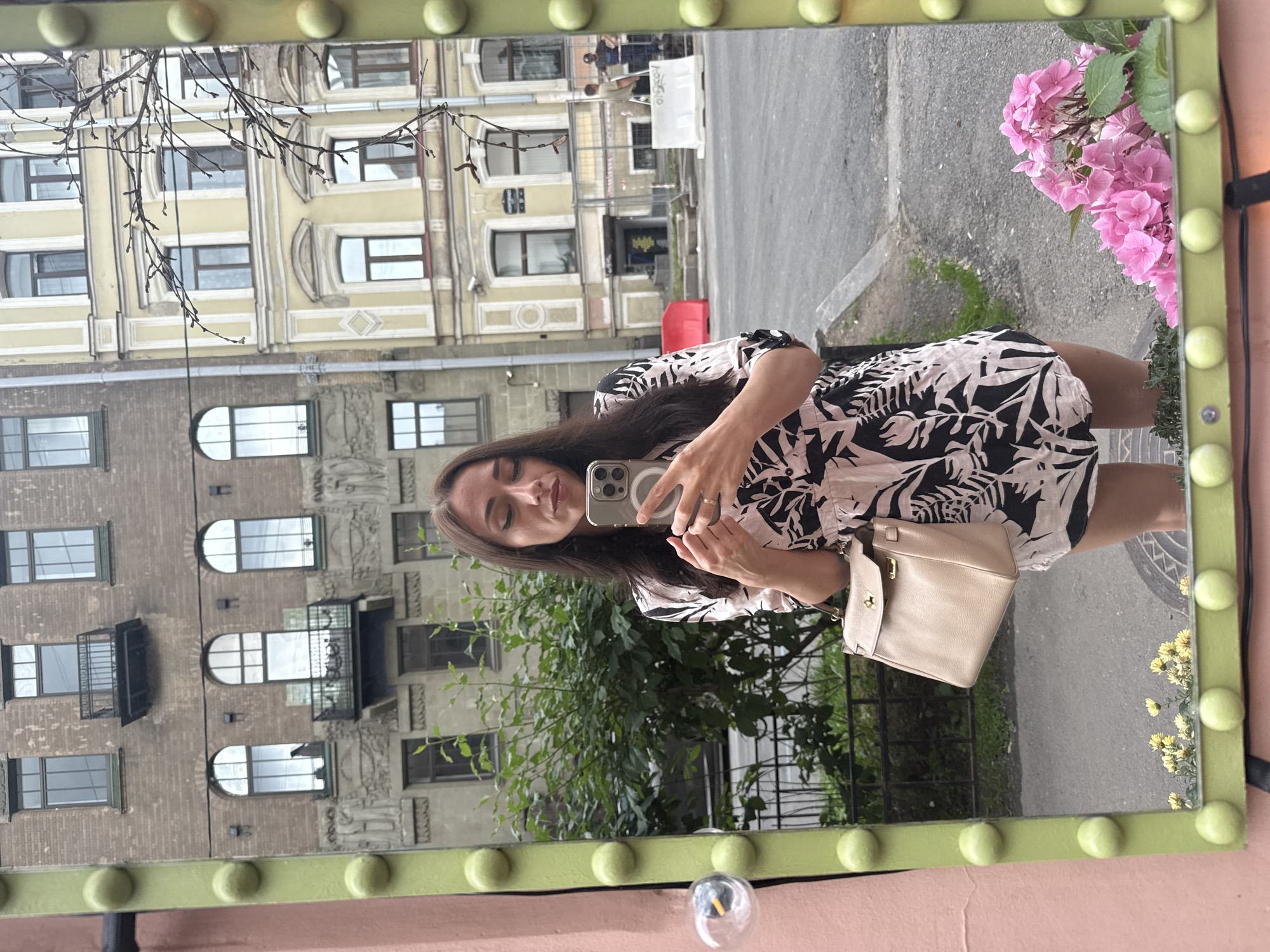 A woman with long dark hair, wearing a black and white floral print dress and holding a beige handbag, is taking a selfie in a large, decorative mirror. The mirror has a lime green frame with protruding green spheres. In the reflection, the woman is looking at her phone, which she is holding up to take the picture. Behind her, reflected in the mirror, are buildings in Saint Petersburg, Russia. One building is made of brown brick with arched windows and balconies. Another building to the right is pale yellow with white decorative molding around its windows and has a sign in Russian. There are some bare tree branches extending into the frame from the right. In the foreground of the reflection, there are small yellow flowers and a cluster of vibrant pink hydrangeas. There are also faint reflections of people and construction barriers across the street. The woman is wearing a ring on her right hand.