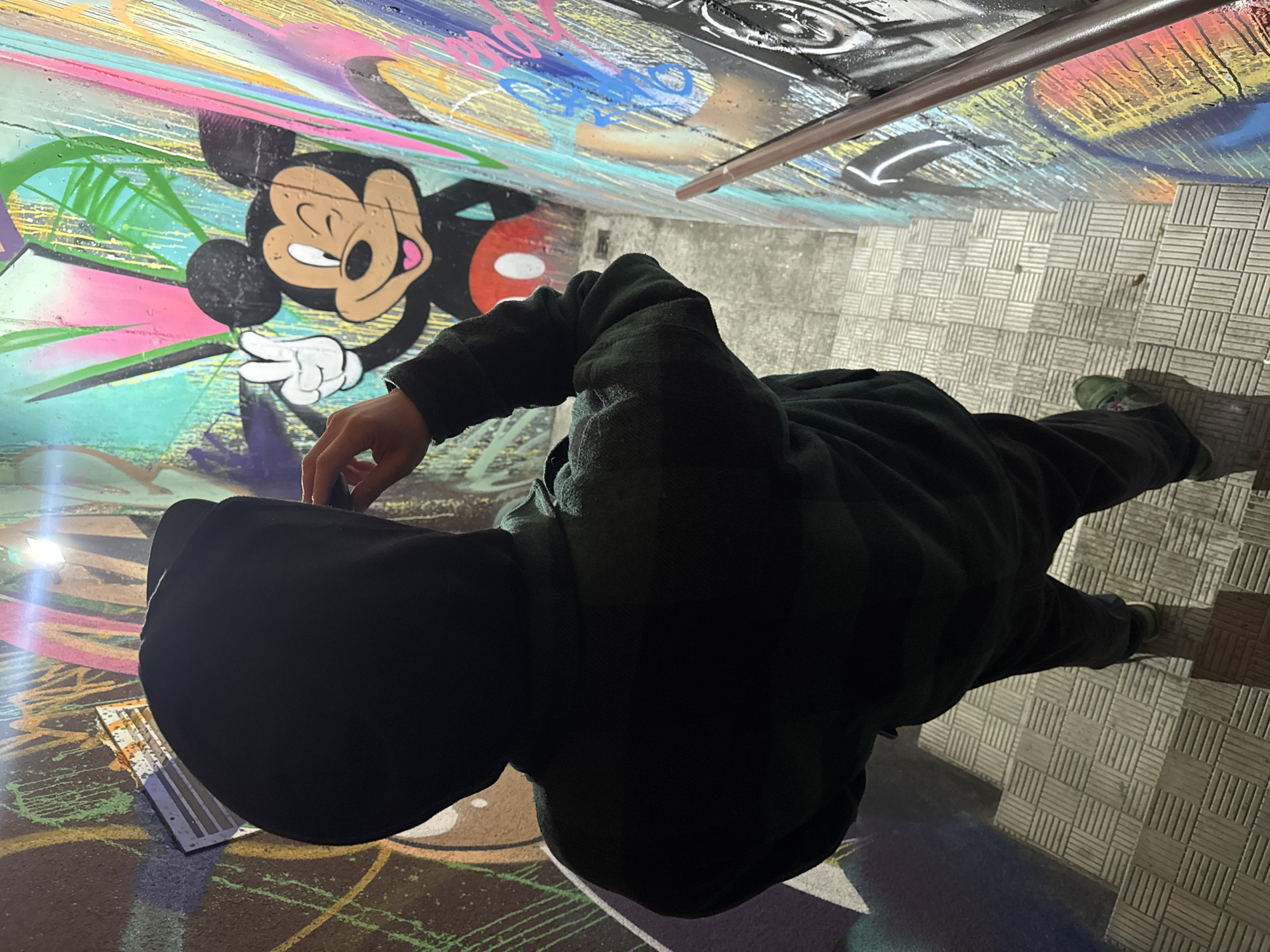 A person in a dark hooded jacket stands in what appears to be an urban underpass or tunnel covered in graffiti in Hrodna, Belarus. The person faces away from the camera, towards a mural depicting Mickey Mouse. The walls and ceiling of the underpass are covered with colorful graffiti art. The floor is a tiled surface. There is a metal bar running along the ceiling. A light source reflects on the wall to the left. The overall lighting is dim.