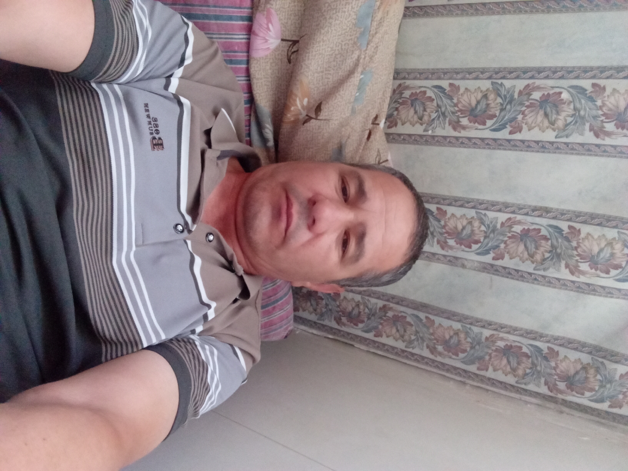 A man is pictured in a selfie, lying down on a couch or bed. He is wearing a collared shirt with horizontal stripes in shades of grey, white, and dark brown. The background features a wall with floral wallpaper in vertical panels. To his left, a pillow with a floral pattern rests behind him. The setting appears to be indoors, likely a domestic environment. There are no explicit indications of Ashgabat, Turkmenistan, or any specific city.
