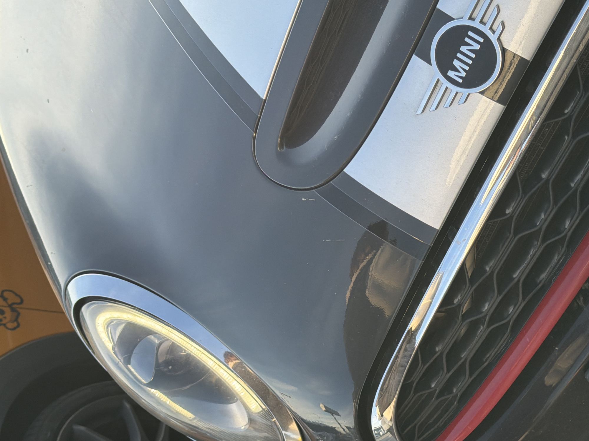 The image presents an angled, close-up view of the front section of a dark-colored Mini Cooper car, observed during what appears to be bright daytime conditions. The car's hood is prominent, featuring a central dark stripe flanked by a lighter, possibly silver, stripe that extends towards the front. A chrome-ringed headlight with an illuminated LED daytime running light is visible in the lower left, casting a yellow glow.
To the right of the hood, a black honeycomb-patterned grille is seen, accented by a horizontal red strip along its lower edge. The car's emblem, a circular "MINI" logo with the brand name in capital letters on a black background surrounded by silver wings, is located on the lighter stripe of the hood.
Reflections of a bright sky and what seem to be environmental elements are visible on the glossy surface of the car's paintwork. Minor scratches and scuffs are noticeable on the dark parts of the hood, and some dirt or residue is present on the lighter stripe near the emblem. Part of a dark car wheel and tire is visible in the bottom left. A yellow object with a cartoon drawing is partially visible on the far left, suggesting proximity to another vehicle or item. The clear reflections and bright lighting indicate sunny weather. While the location context of Saint Petersburg, Russia, is provided, there are no visual cues within the image to confirm this specific geographical location.