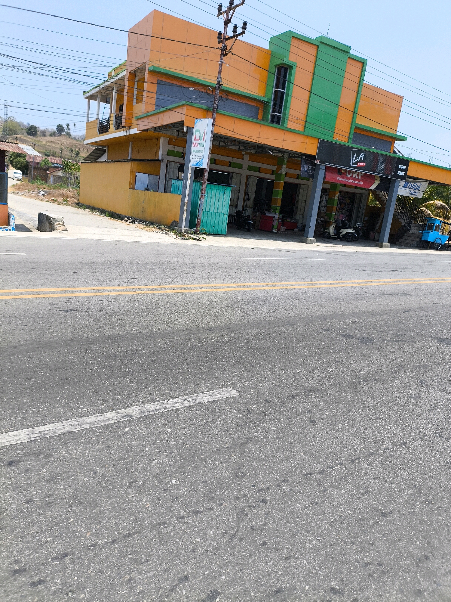 The image captures a street view in Kwandang, Indonesia, featuring a prominent orange and green building. The building has multiple levels, balconies, and storefronts at the ground level. Signs are visible on the building, including "LA COLOR" and "DI. DRP". Several motorbikes are parked in front of the storefronts. A blue street cart is parked to the right of the building. The street in the foreground is asphalt with yellow and white lane markings. A rocky embankment is visible behind the building, with a few trees and houses in the distance. Overhead power lines crisscross the sky, which is clear and blue. The scene appears to be a typical day in a bustling commercial area.
